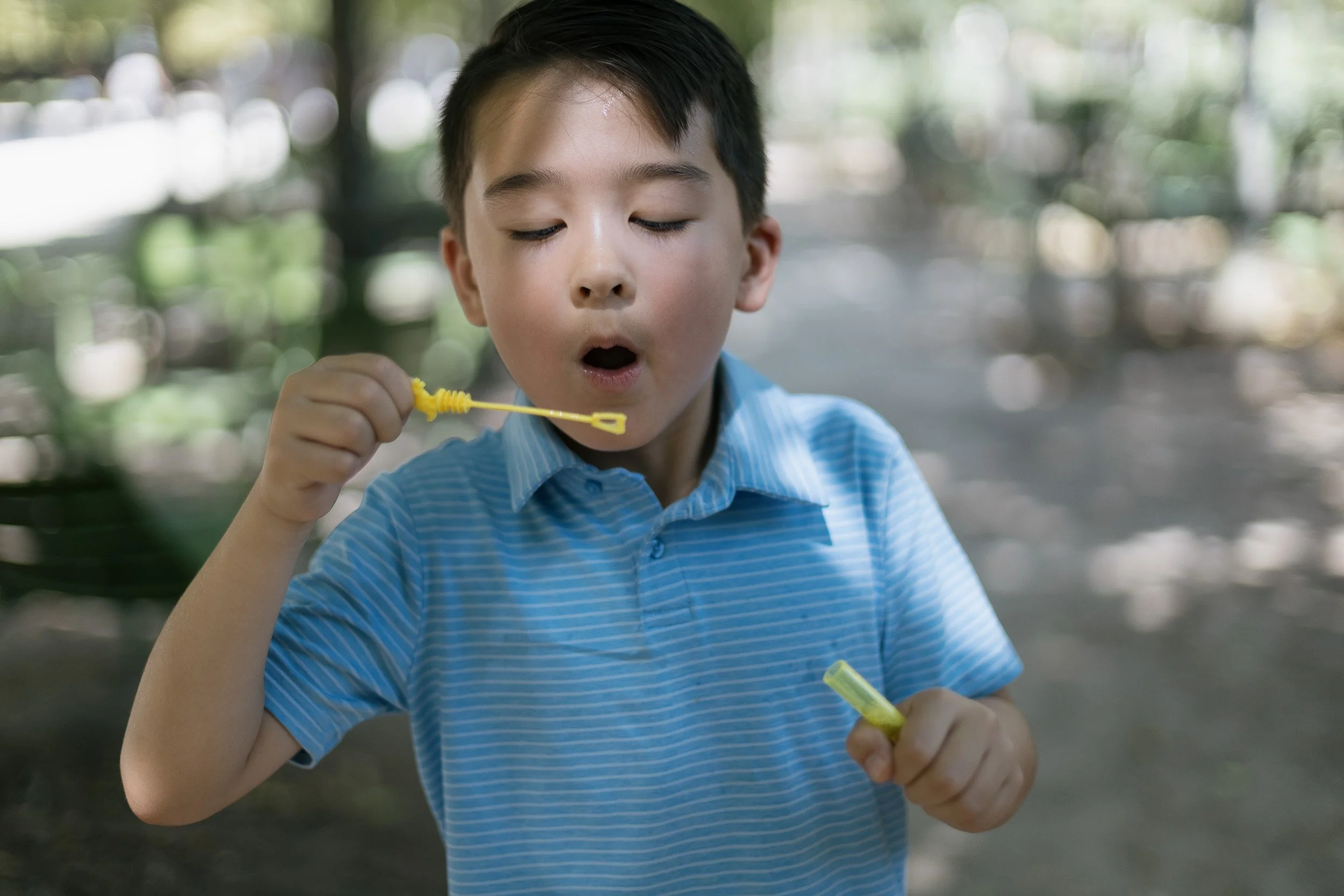 Young boy with wet hair, wearing a blue and white striped shirt, is blowing bubbles outdoors in a park with blurred trees and natural background photography by Maria Zvir.