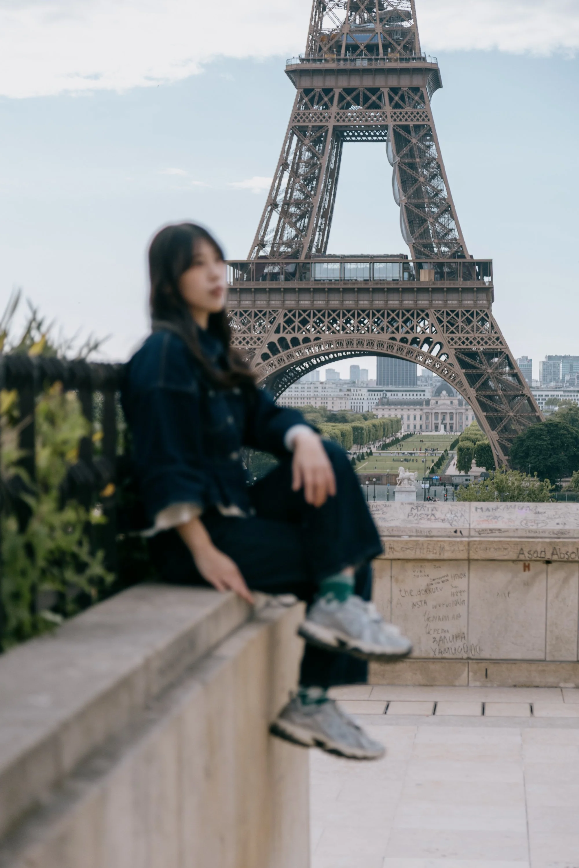 A woman sitting on a ledge in front of the Eiffel Tower in Paris, France, with graffiti on the wall below.
