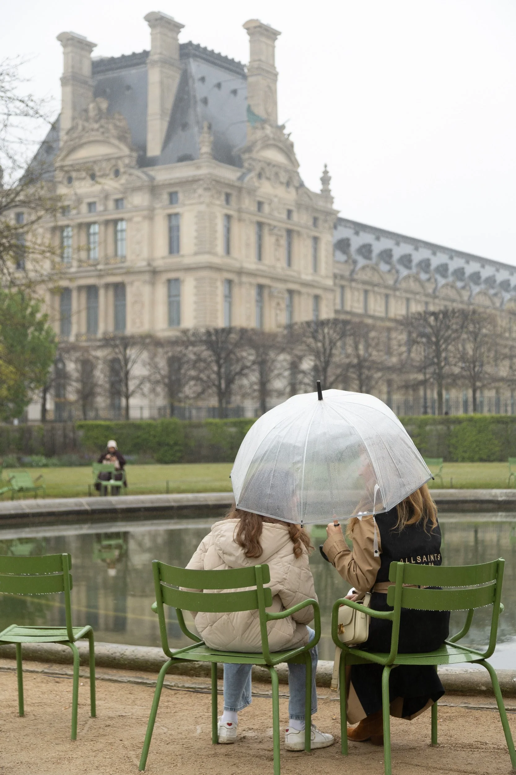 Two women sitting on green chairs by a pond, holding an umbrella in rainy weather. In the backdrop, there is a large historic building, trees, and a person sitting on a bench.