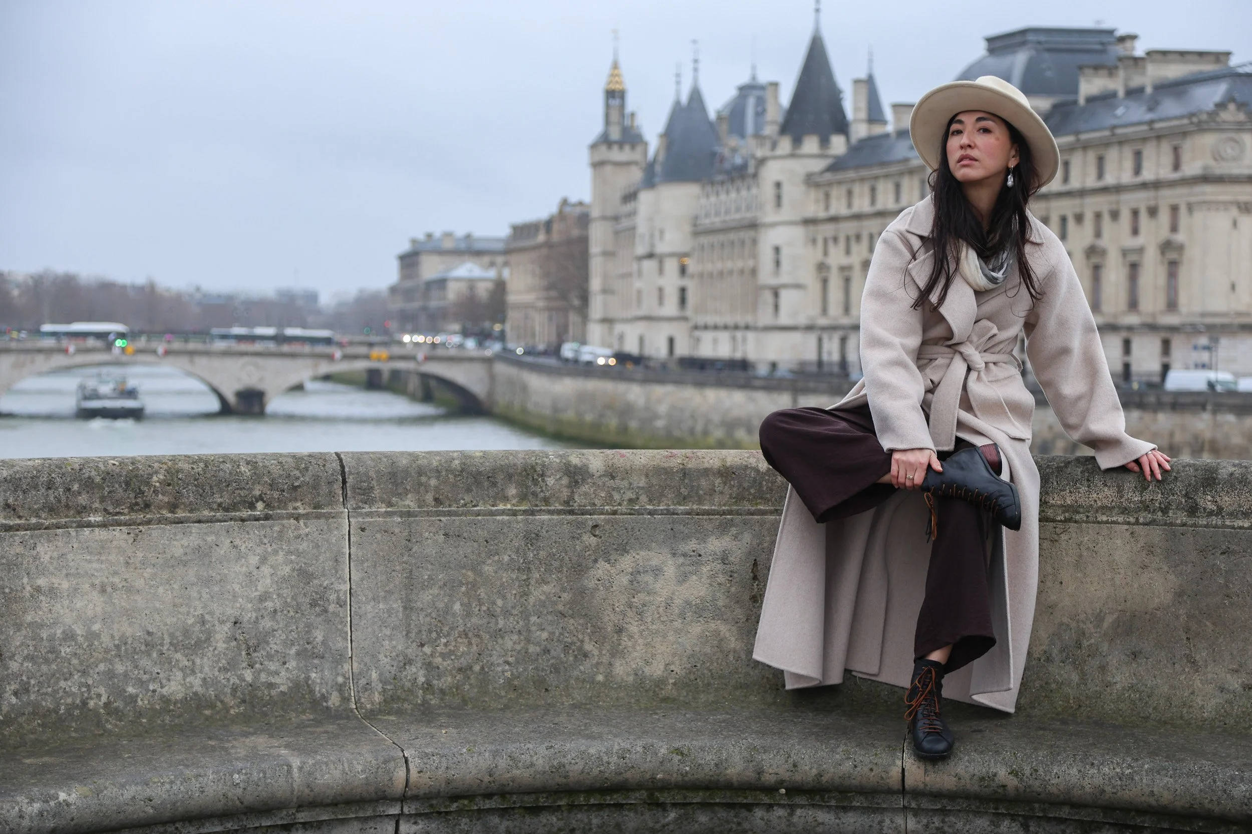 A woman sitting on a stone ledge with a castle-like building and a bridge over a river in the background.