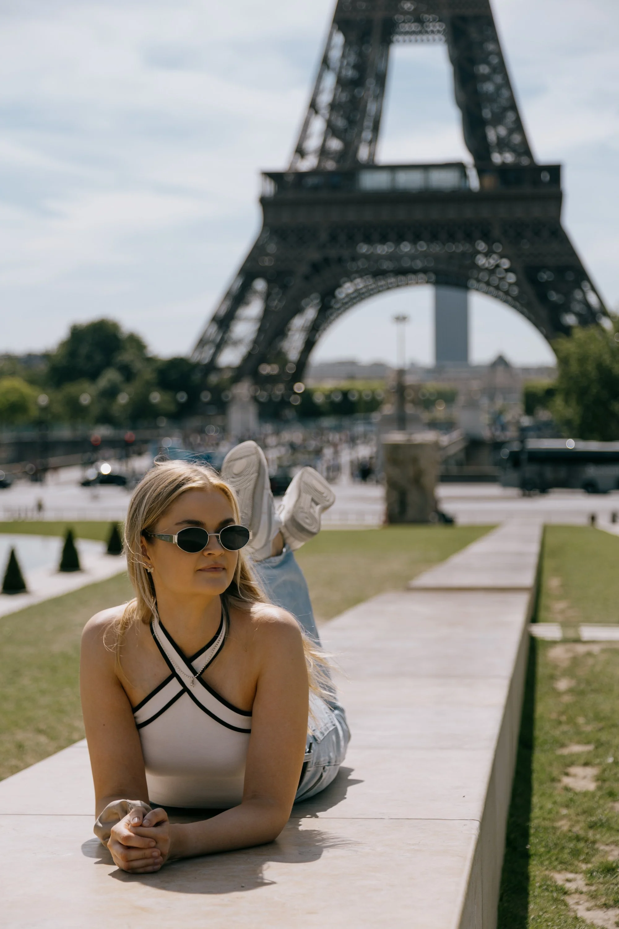 A woman with sunglasses lying on her stomach on a wooden bench outside, with the Eiffel Tower in Paris visible in the background.