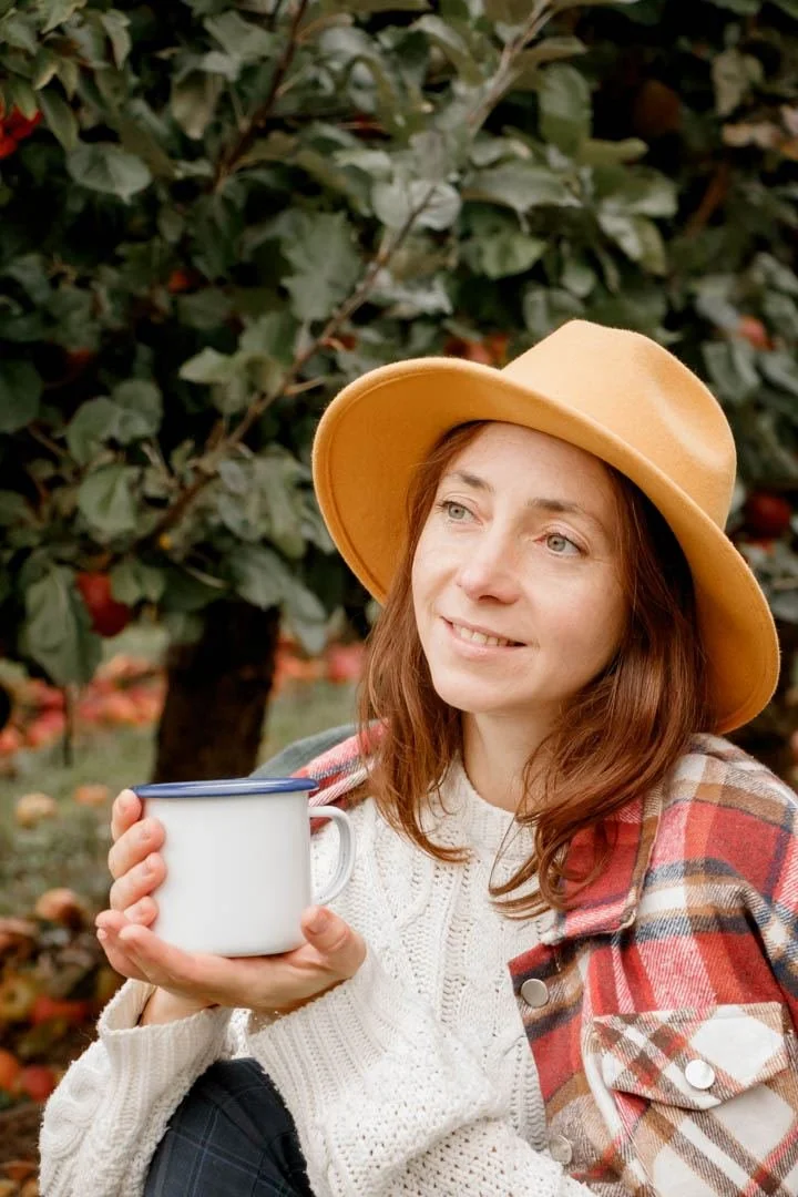 A woman wearing a beige hat and a plaid jacket sitting outdoors among apple trees, holding a white mug and smiling.