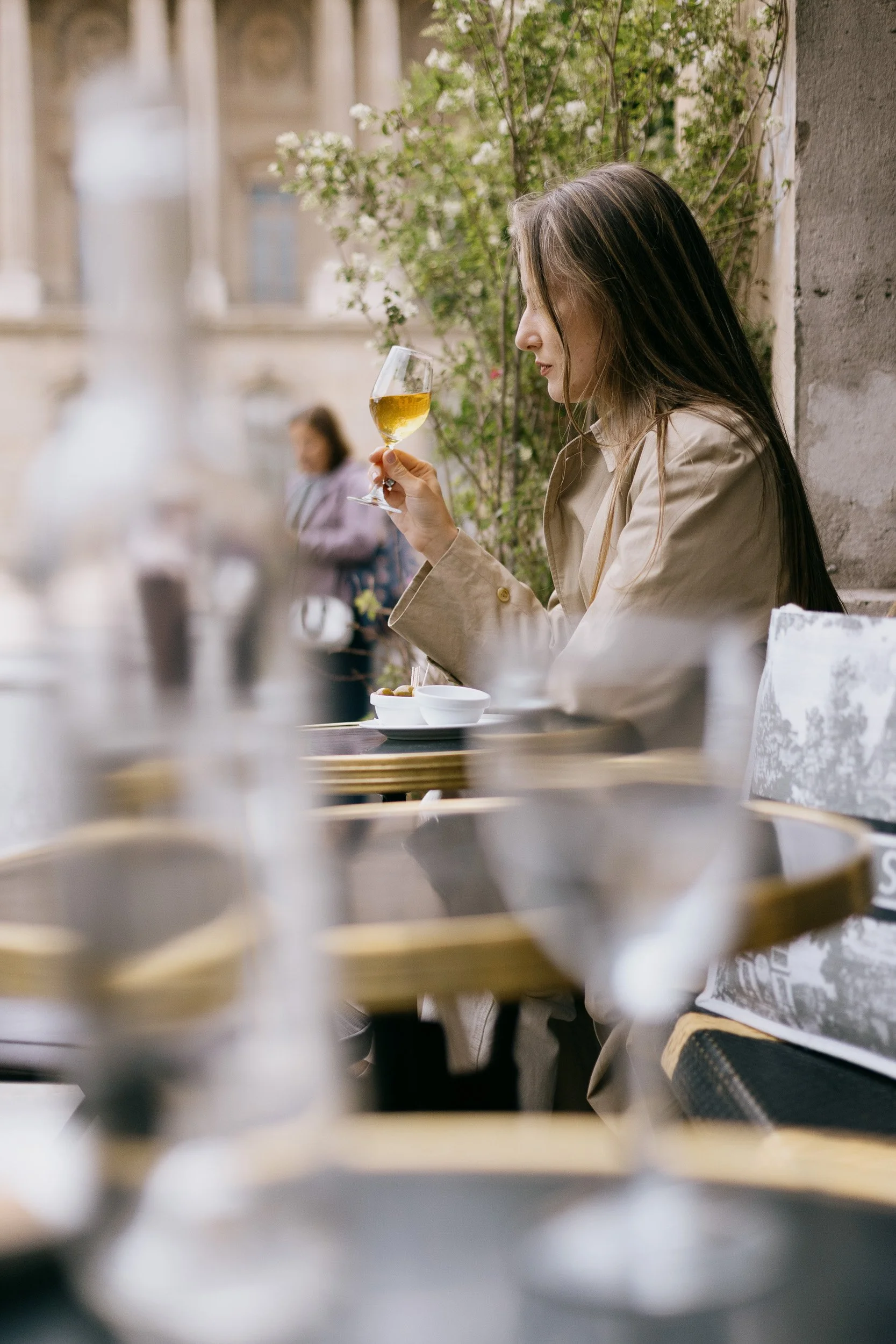 A woman with long brown hair sitting outdoors, holding a glass of white wine, with a blurred background of greenery and a building, and a woman walking in the distance.