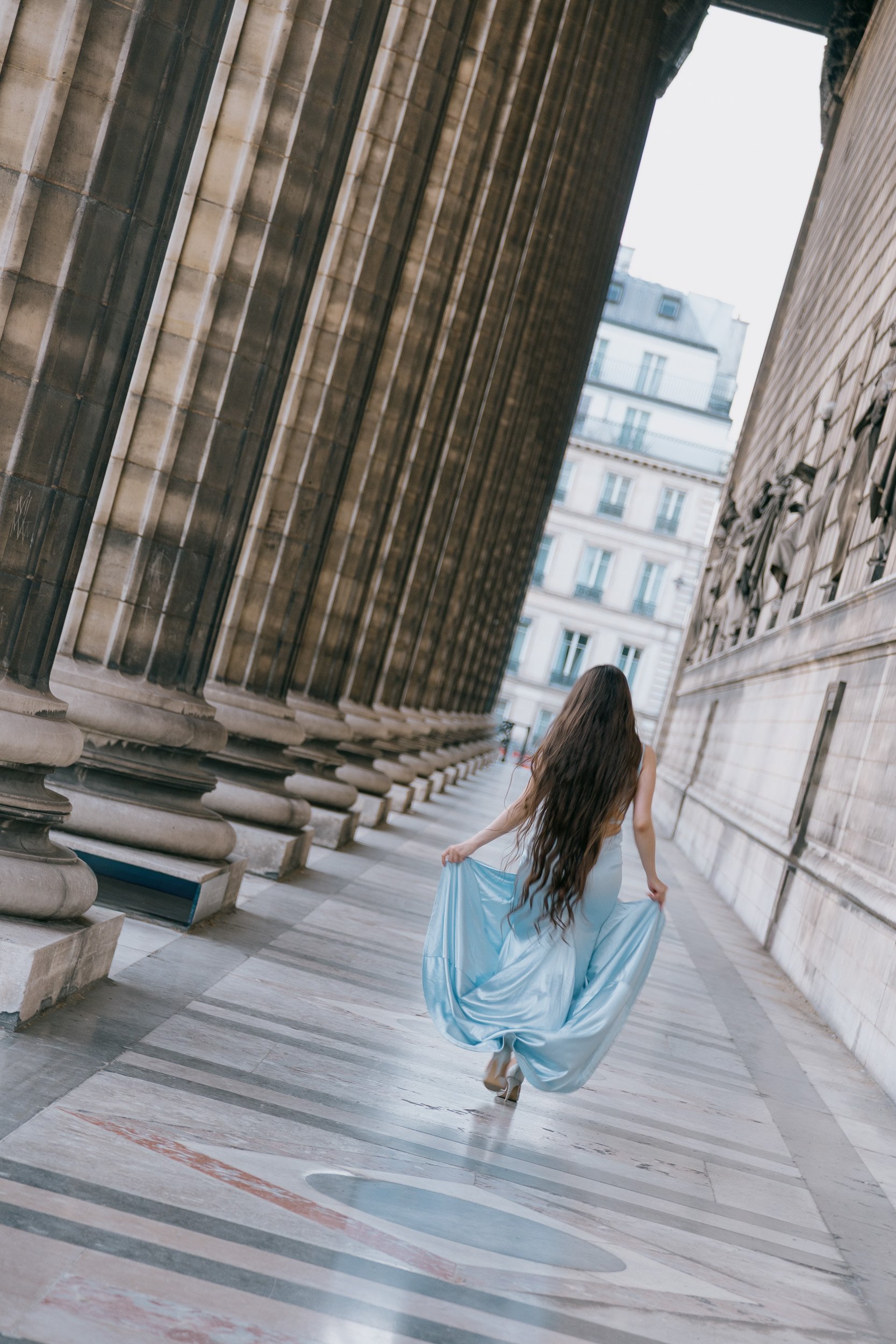 A woman with long flowing hair wearing a light blue gown, running through a corridor between historic building facades in a city.