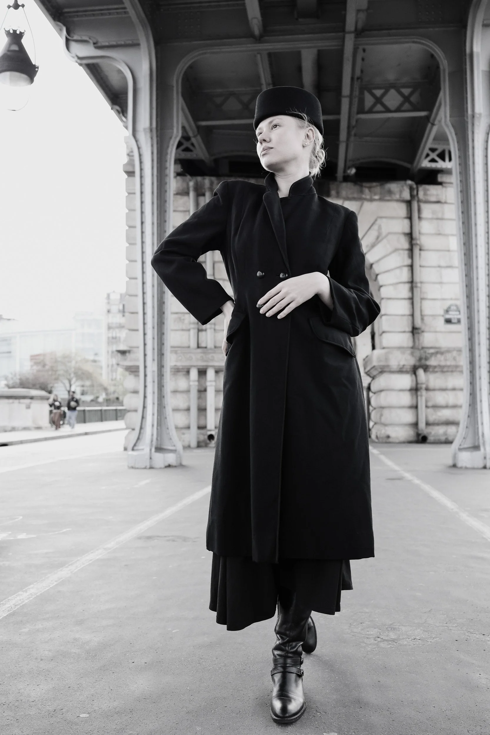 A woman wearing a black coat, black hat, and black boots walking under a bridge in black and white.