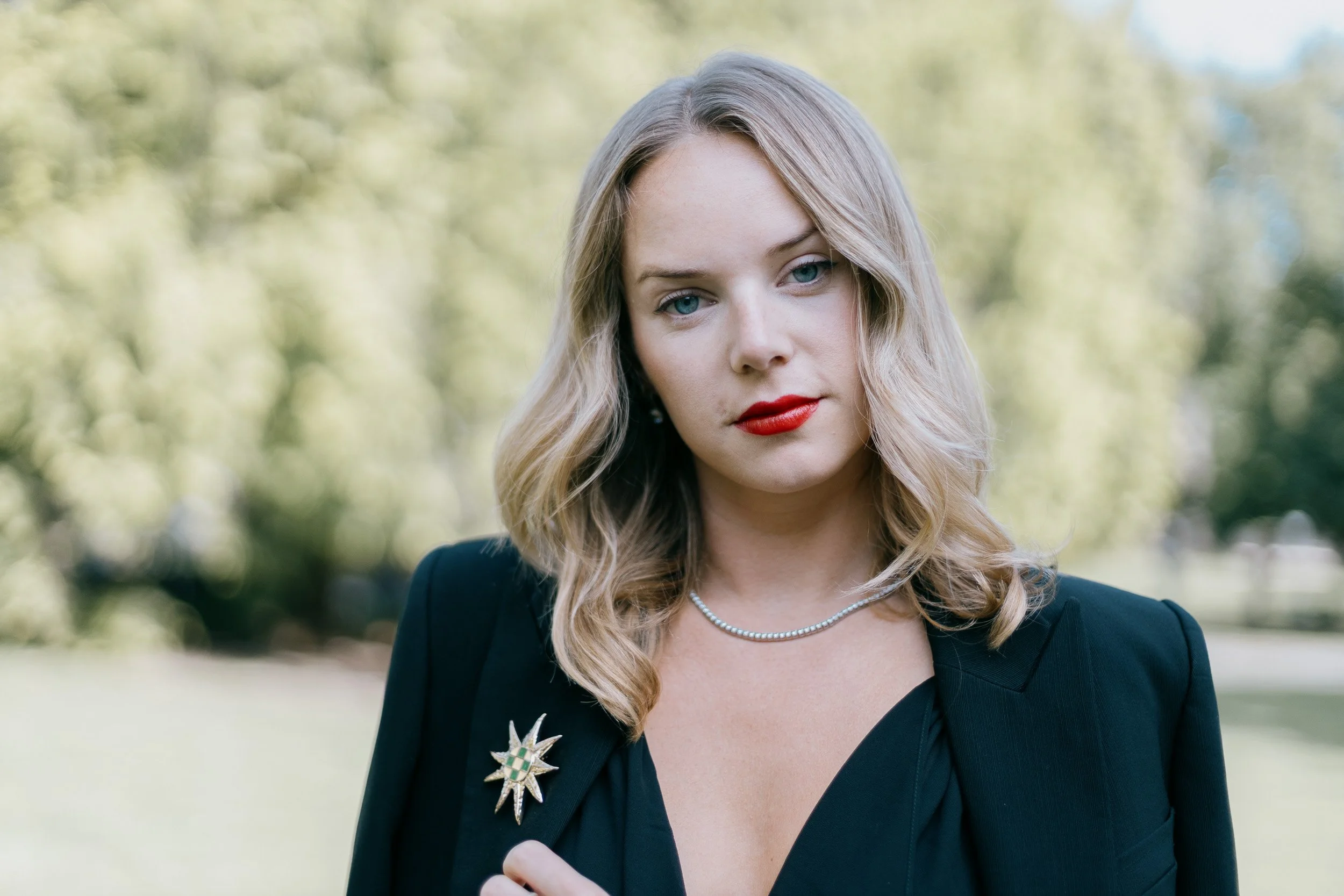 A woman with blonde hair, blue eyes, red lipstick, wearing a black jacket with a star-shaped brooch, and a pearl necklace, standing outdoors with a blurred green and yellow background.