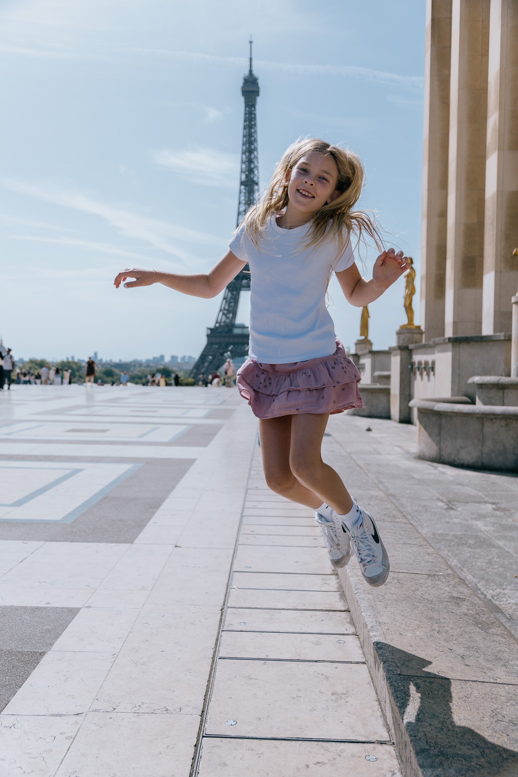 A young girl jumping on a sidewalk in front of the Eiffel Tower in Paris, France, during the daytime.