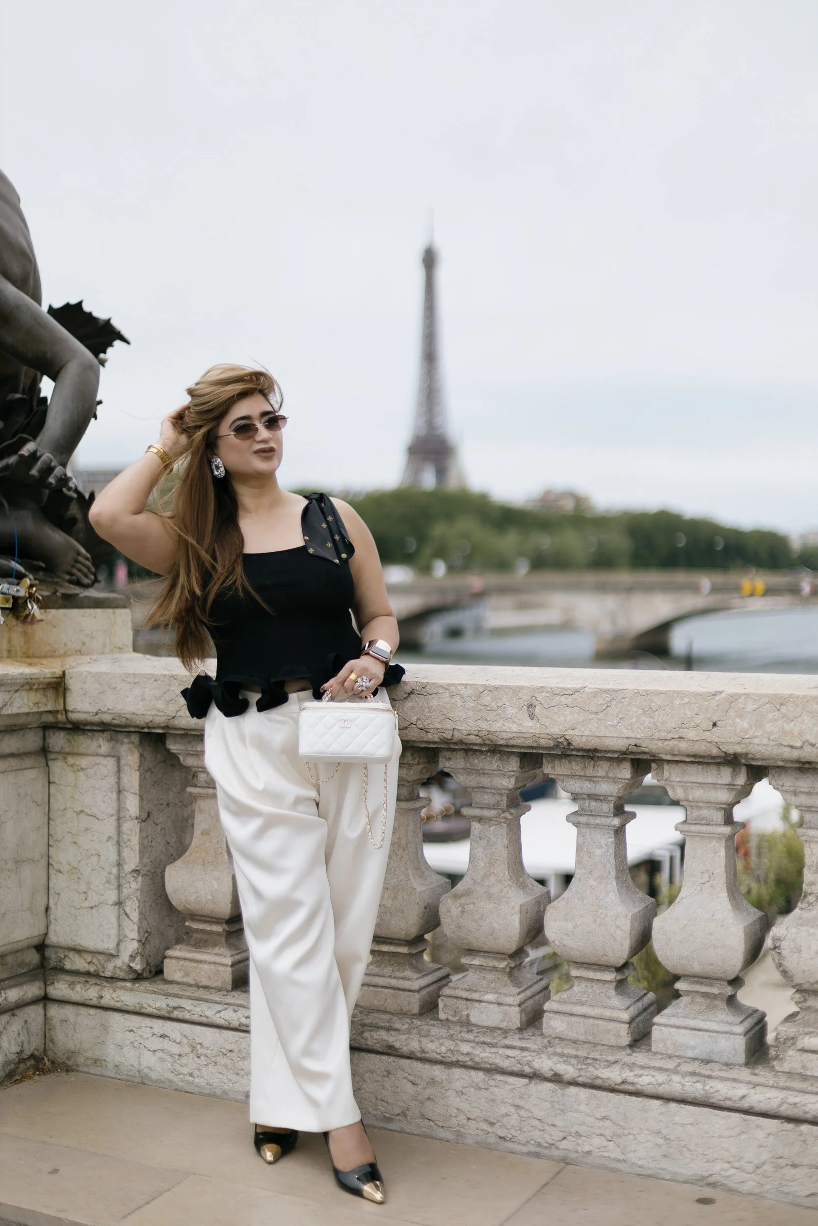 A woman with long brown hair, sunglasses, wearing a black sleeveless top with ruffle details, white high-waisted pants, and black shoes with gold tips, posing on a stone balcony in front of a railing with the Eiffel Tower in the background in Paris, France.
