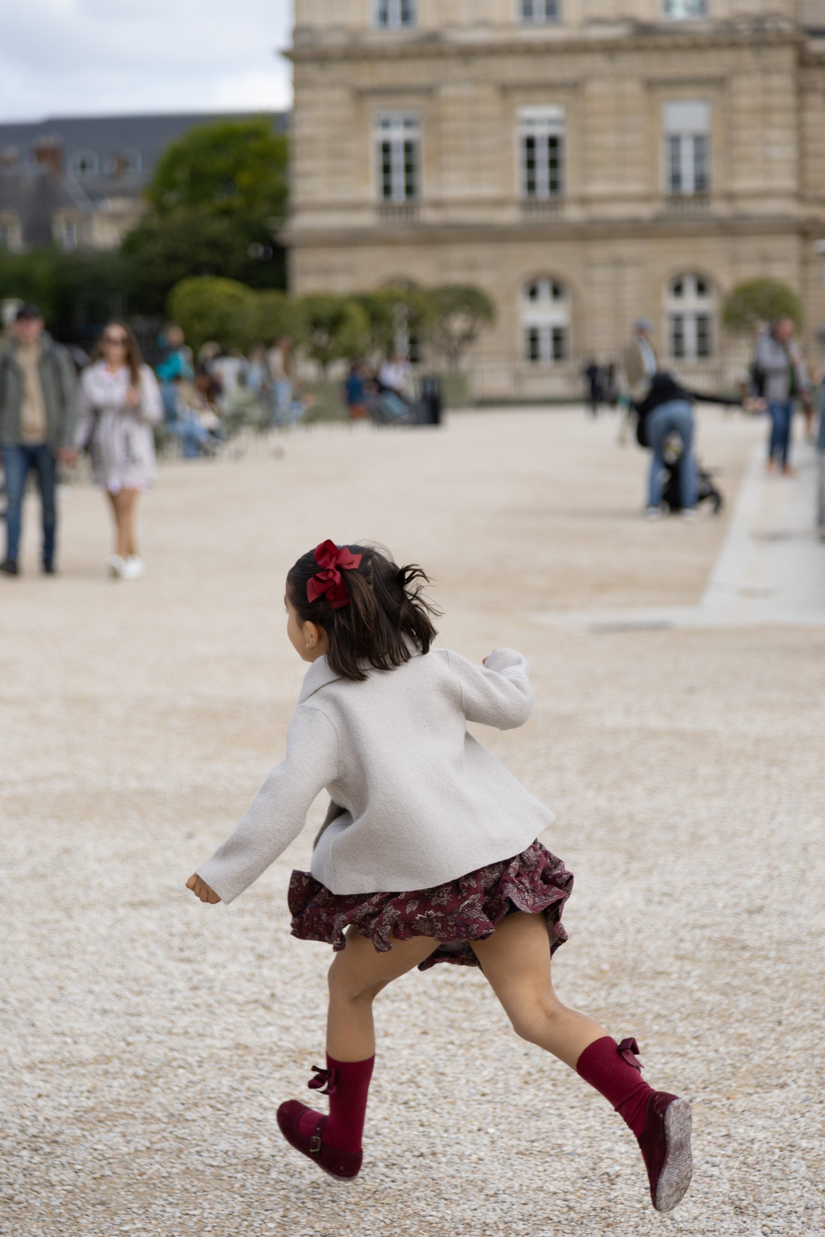 Young girl with a red bow in her hair playing in a gravel area, with a historic building and other people in the background.