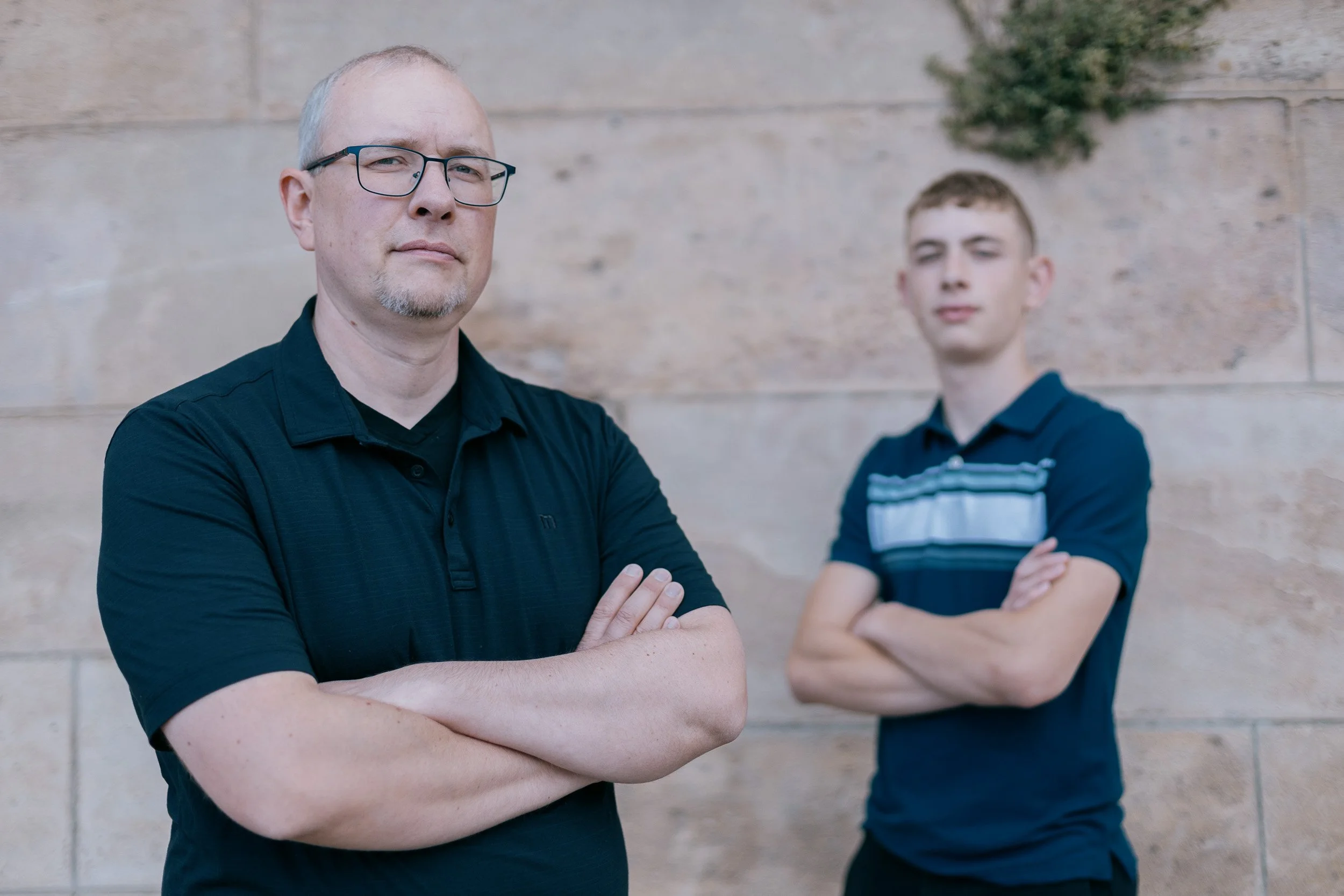 A man with glasses and a goatee standing with his arms crossed in front of a brick wall, with a young boy with curly hair and arms crossed in the background.