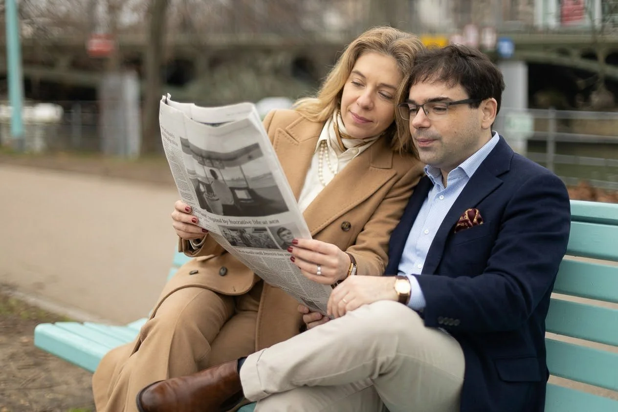 A woman and a man sitting on a park bench, reading a newspaper together.