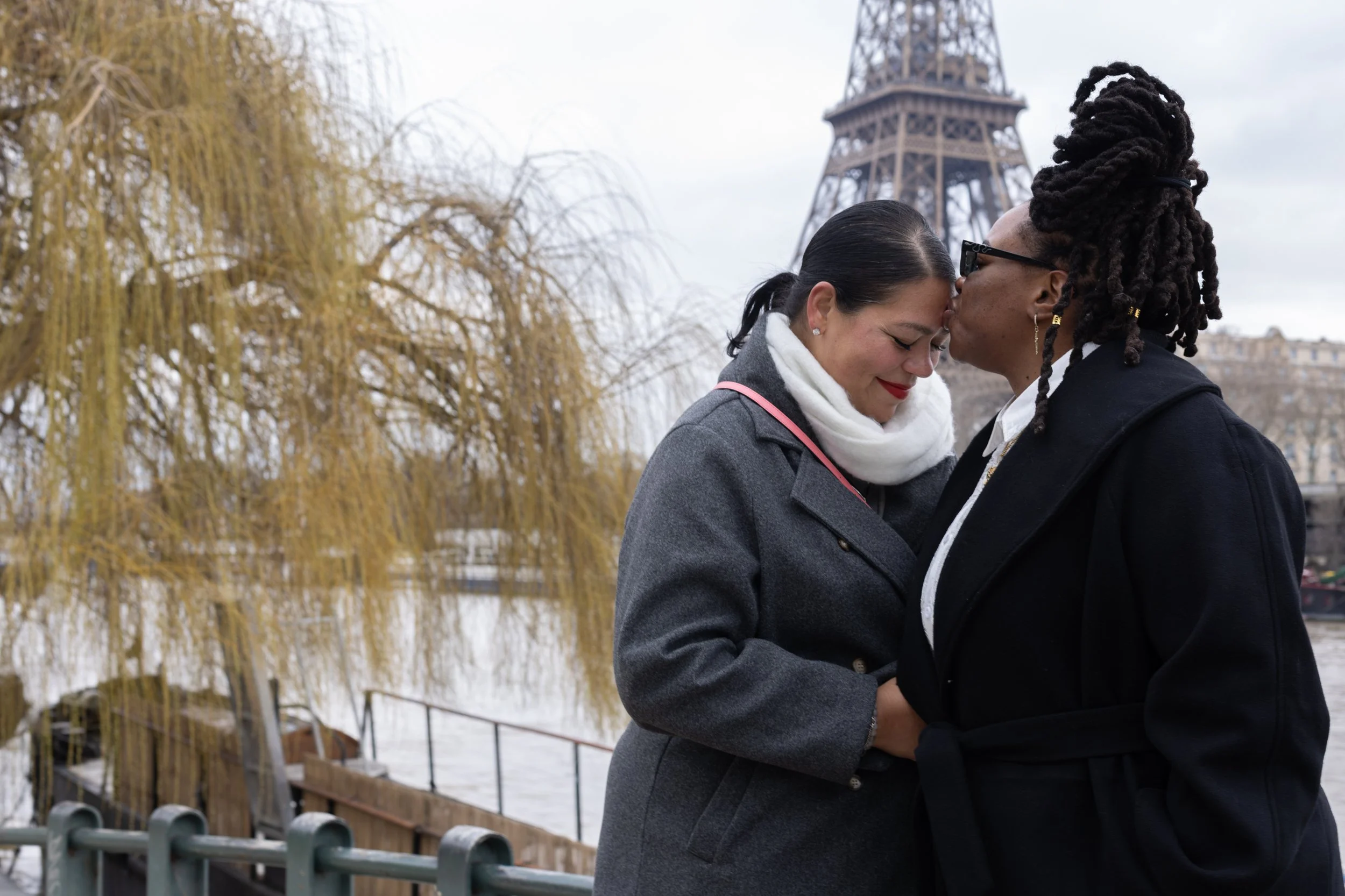 Two women are standing close together, touching foreheads affectionately. One woman has dark hair in a ponytail and is wearing a gray coat with a white scarf, while the other has dreadlocks and glasses, wearing a black coat. The Eiffel Tower is visible in the background along with some trees and a cloudy sky.