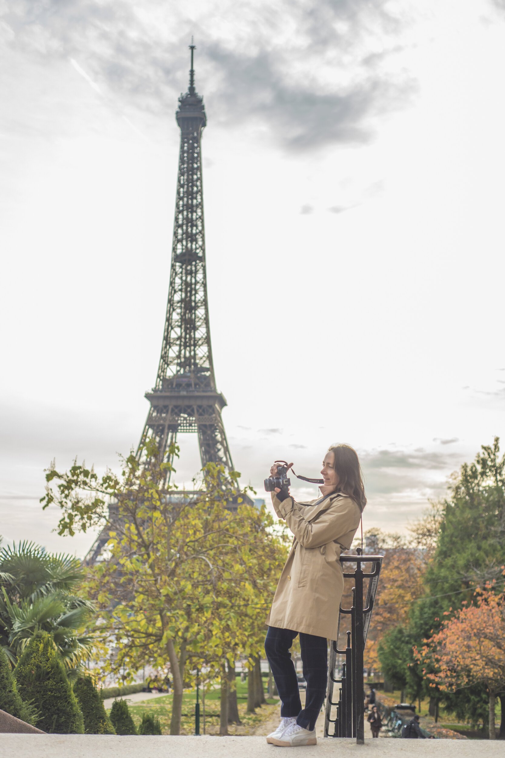 A woman in a beige coat and white sneakers takes a photo with her camera at the base of the Eiffel Tower in a park with trees showing fall foliage.