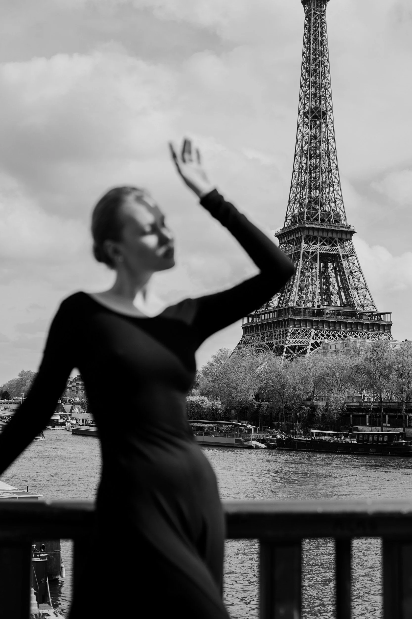 A woman with short hair is standing near a river during the day, with her hand raised in front of her face. In the background, the Eiffel Tower is visible under a cloudy sky.
