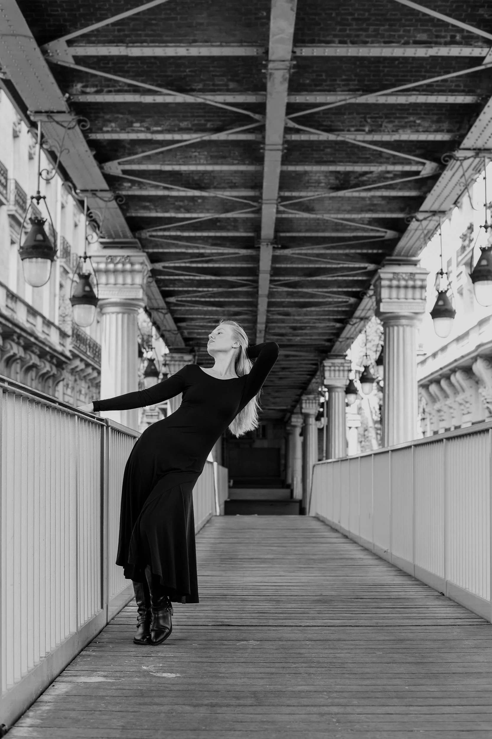 A woman in a long black dress and boots is posing on a bridge with a metal railing, under a bridge or overpass with decorative columns and vintage-style lamps, black and white photo.