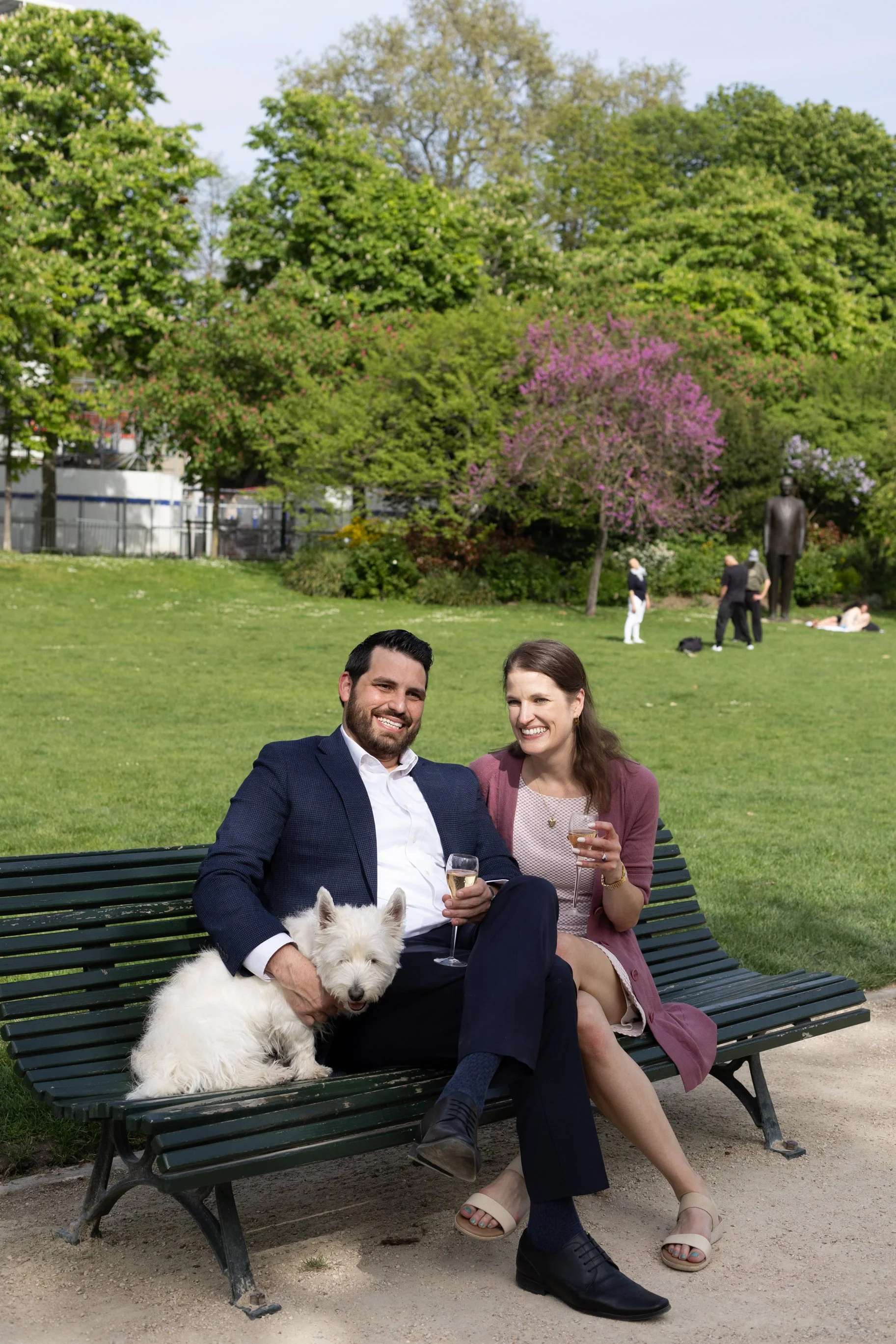 A man and a woman sitting on a park bench, smiling, with a white dog between them, holding a glass of champagne in each hand. The scene is set in a green park with trees and people in the background.