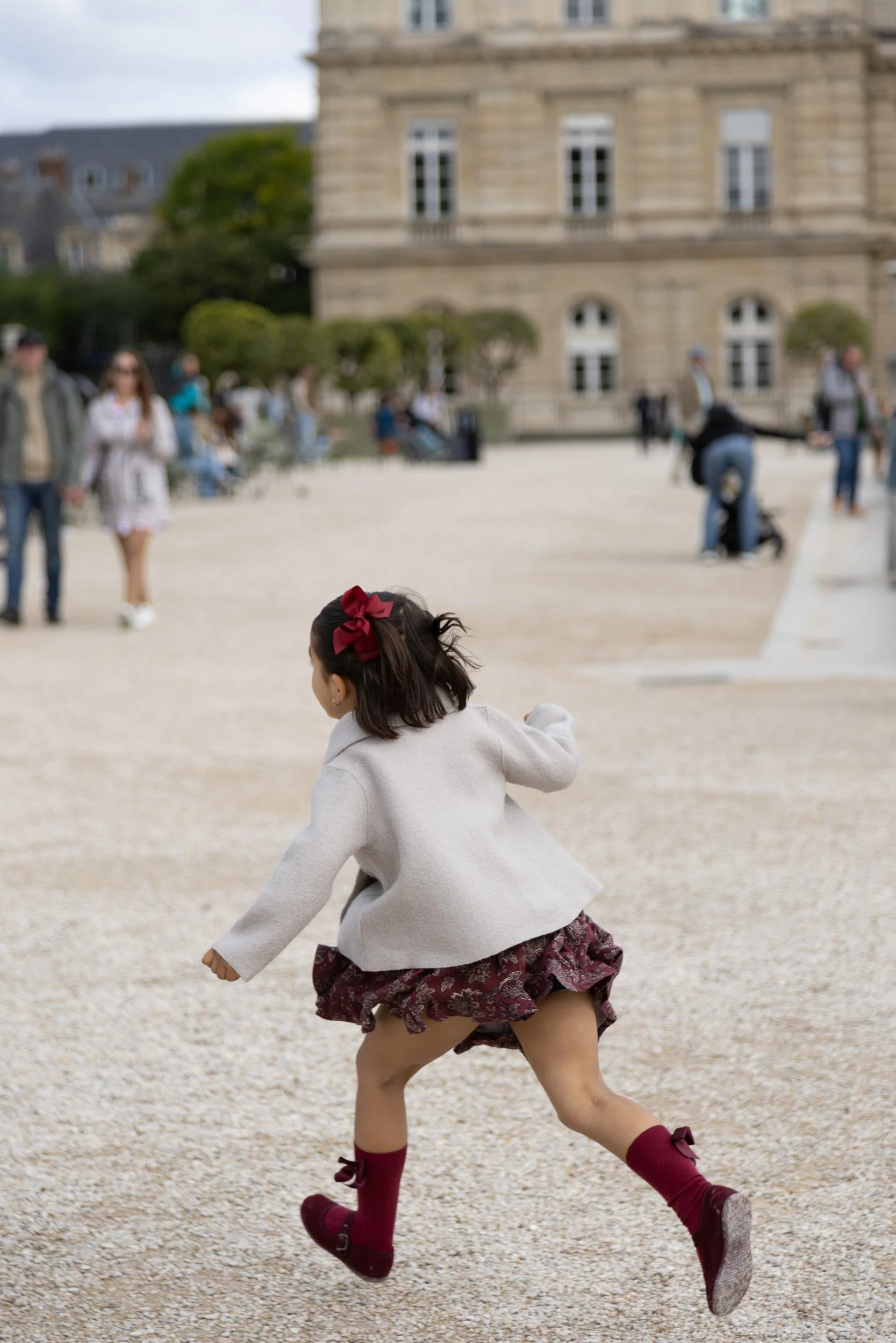 A young girl with a red bow in her hair running on a gravel surface in front of a historic building with people walking and sitting in the background.