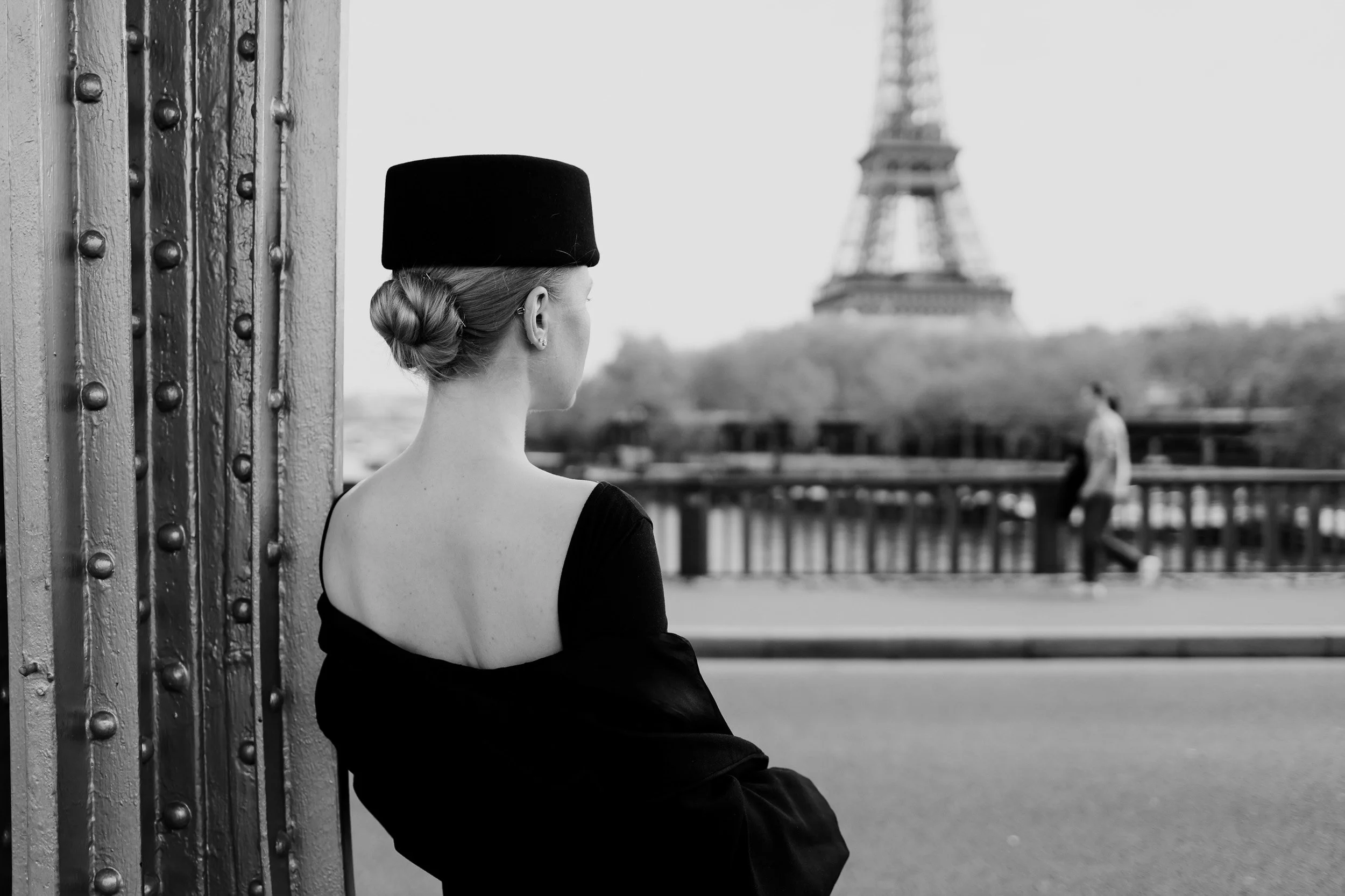 A woman in black ballet attire with her hair in a bun, standing near a metal structure, looking at the Eiffel Tower in the distance, in black and white.