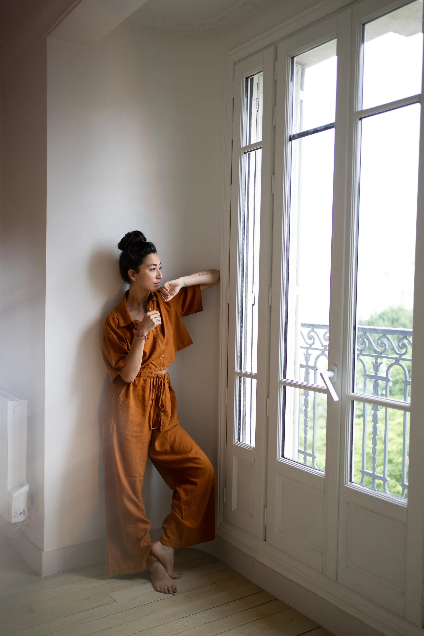 A woman dressed in brown pants and a matching top stands near a large window, with her back against a white wall, looking outside.