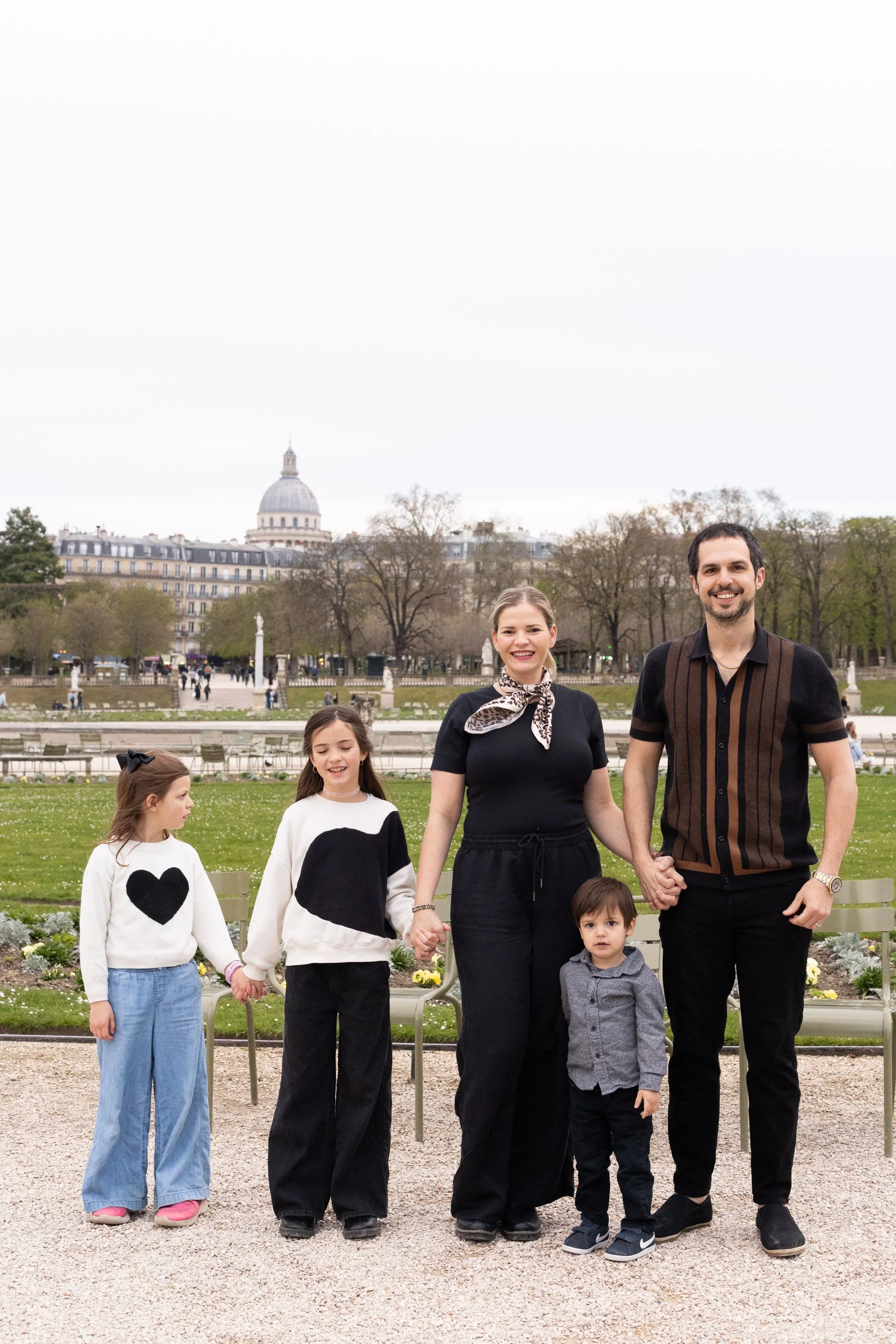 A family of seven holding hands and smiling outdoors in front of a park with trees and a cityscape with a notable dome building in the background.