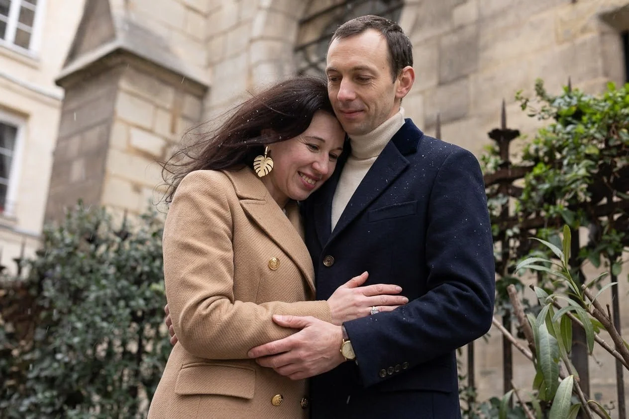 A couple embracing outdoors near a stone building, smiling with their heads touching, wearing elegant coats; the woman has dark hair and gold leaf earrings, the man has short hair and a watch.