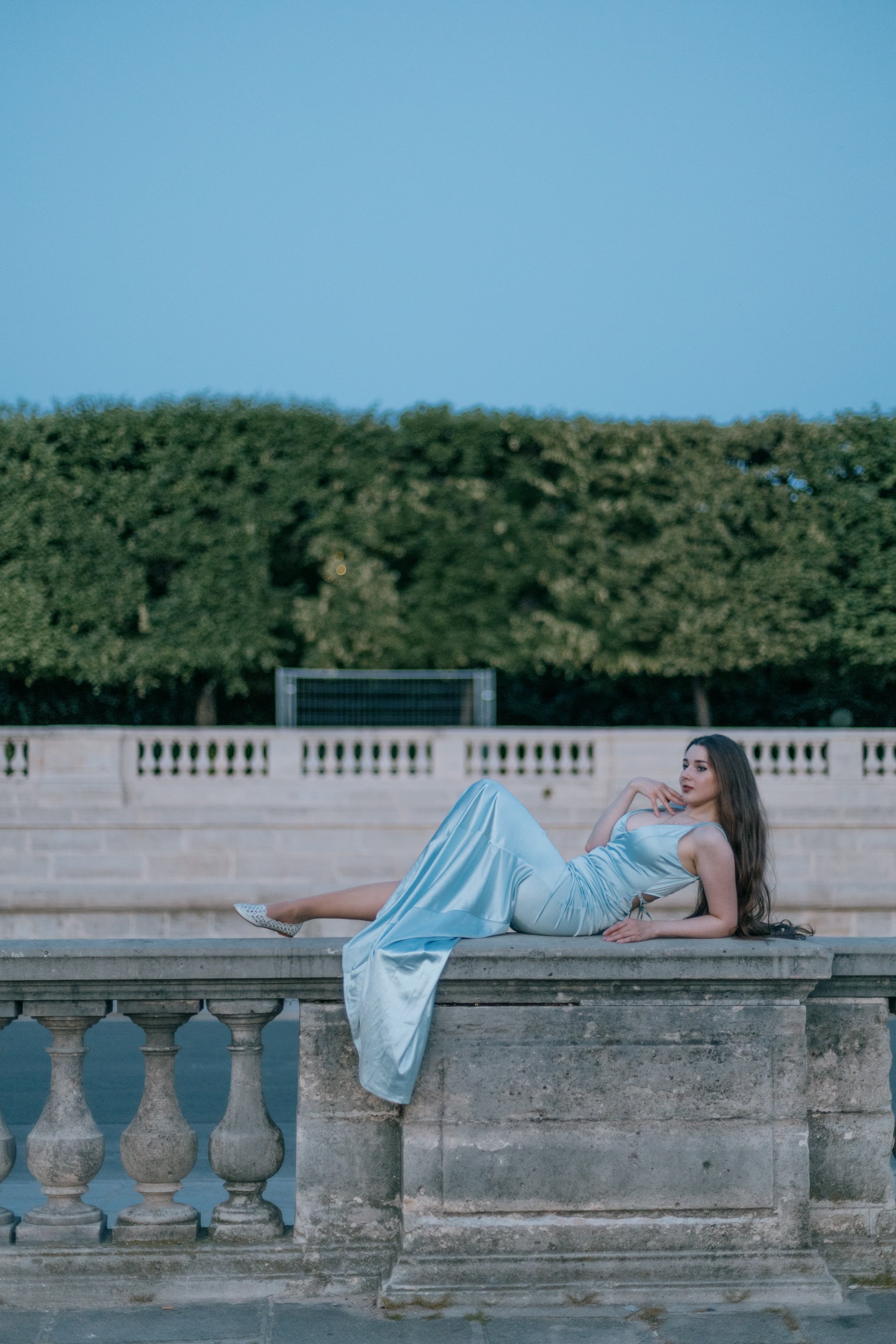 A woman in a light blue satin dress reclining on a stone ledge with a balustrade, a large hedge, and a blue sky in the background.