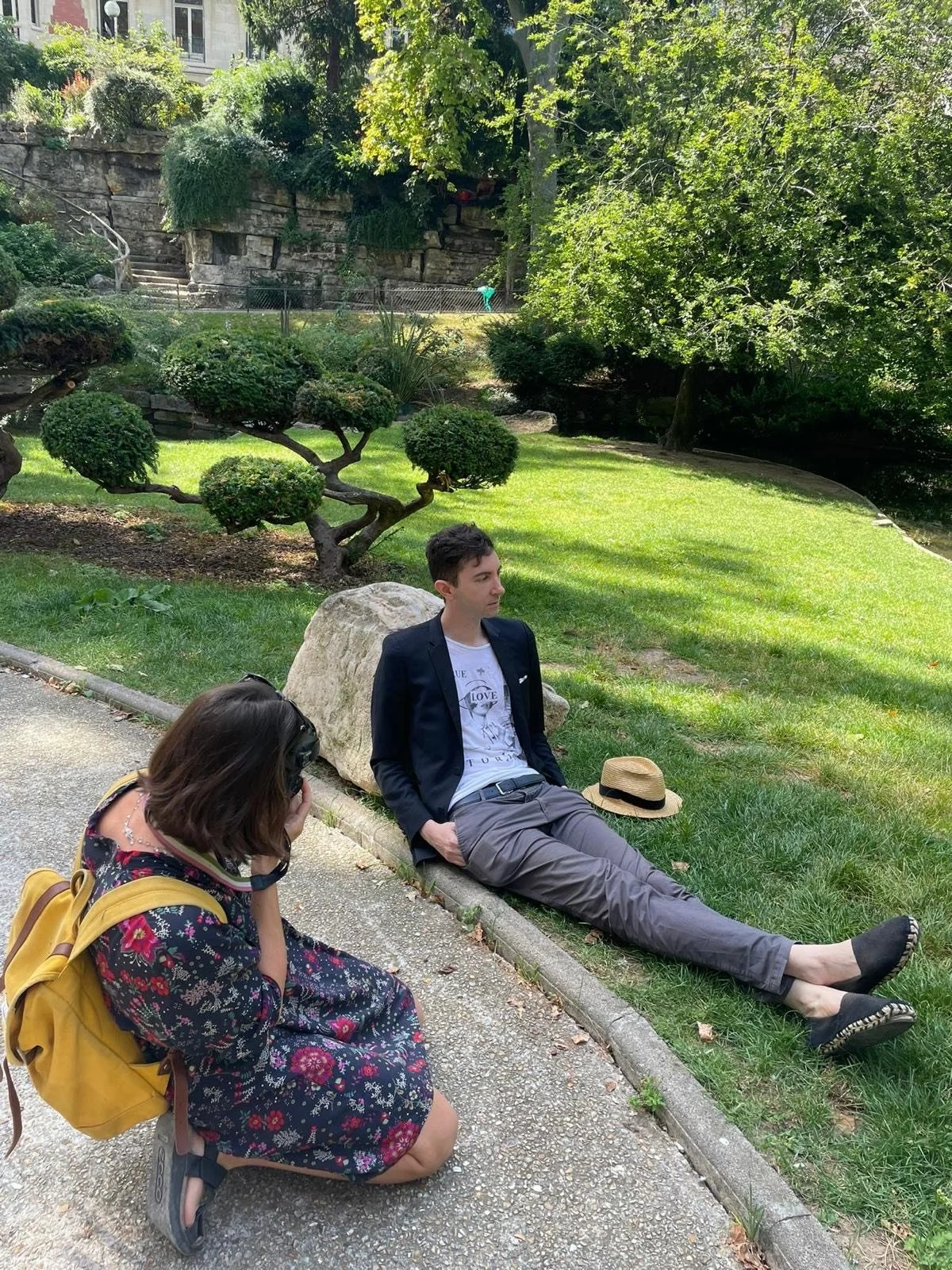 A young woman with a yellow backpack kneels on the ground taking a picture of a young man sitting on the grass in a park. The young man is wearing a suit jacket, white t-shirt, gray pants, and black shoes, with a hat placed on the grass beside him. The park has well-maintained green grass, decorative trees, and dense foliage in the background.