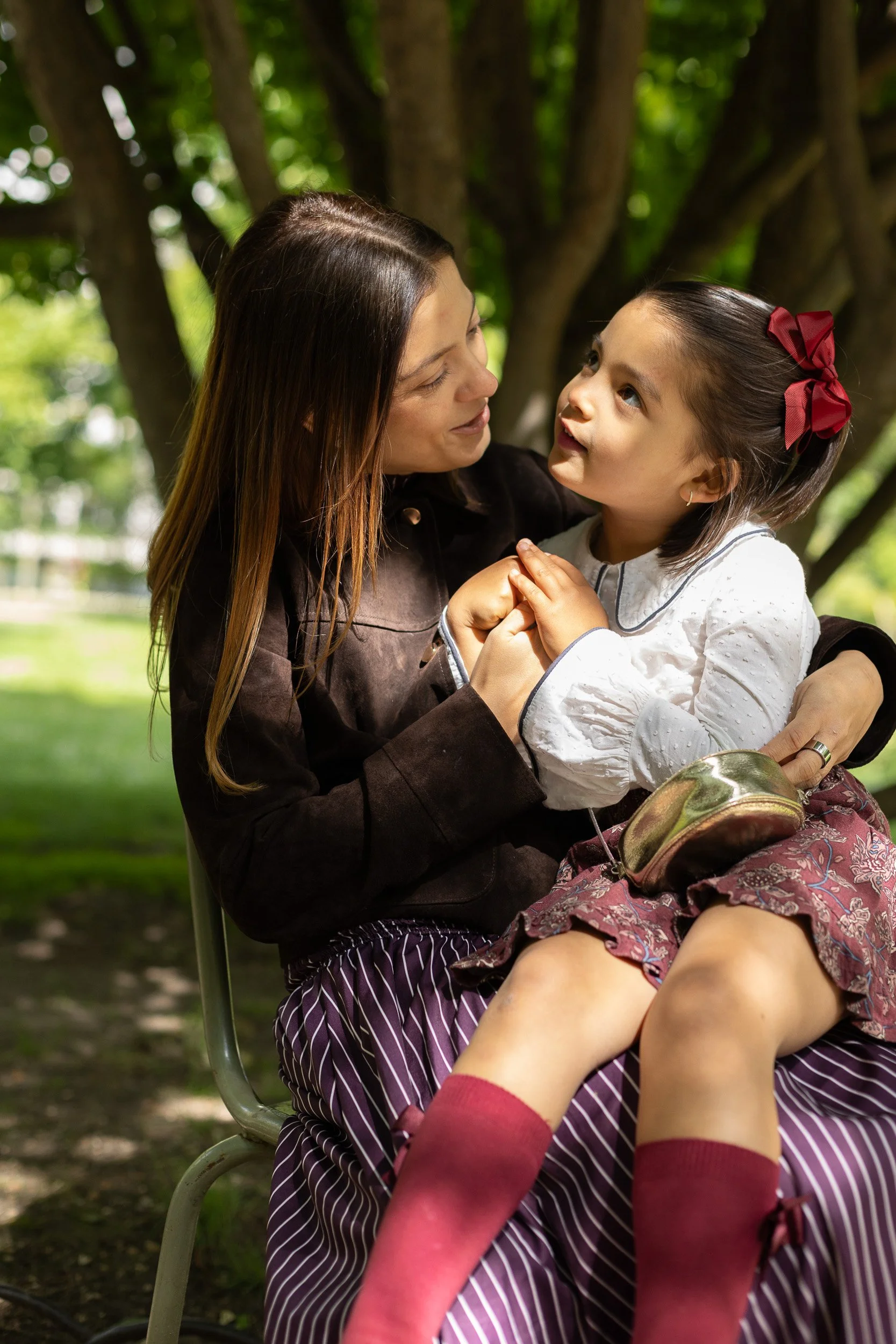 A woman and a young girl are sitting outdoors under a tree, engaging in a conversation. The woman has long brown hair and is wearing a dark jacket, while the girl has brown hair tied with a large red bow, and is wearing a white shirt with black trim and a patterned skirt. The girl is sitting on a chair with her feet raised, wearing red socks and shoes.