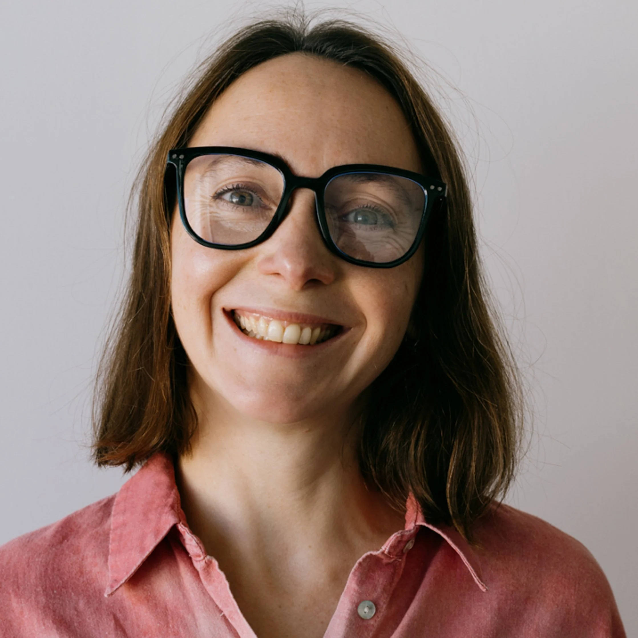 Close-up of a smiling woman with light skin, brown hair, and black glasses, wearing a pinkish-red collared shirt against a plain white background.