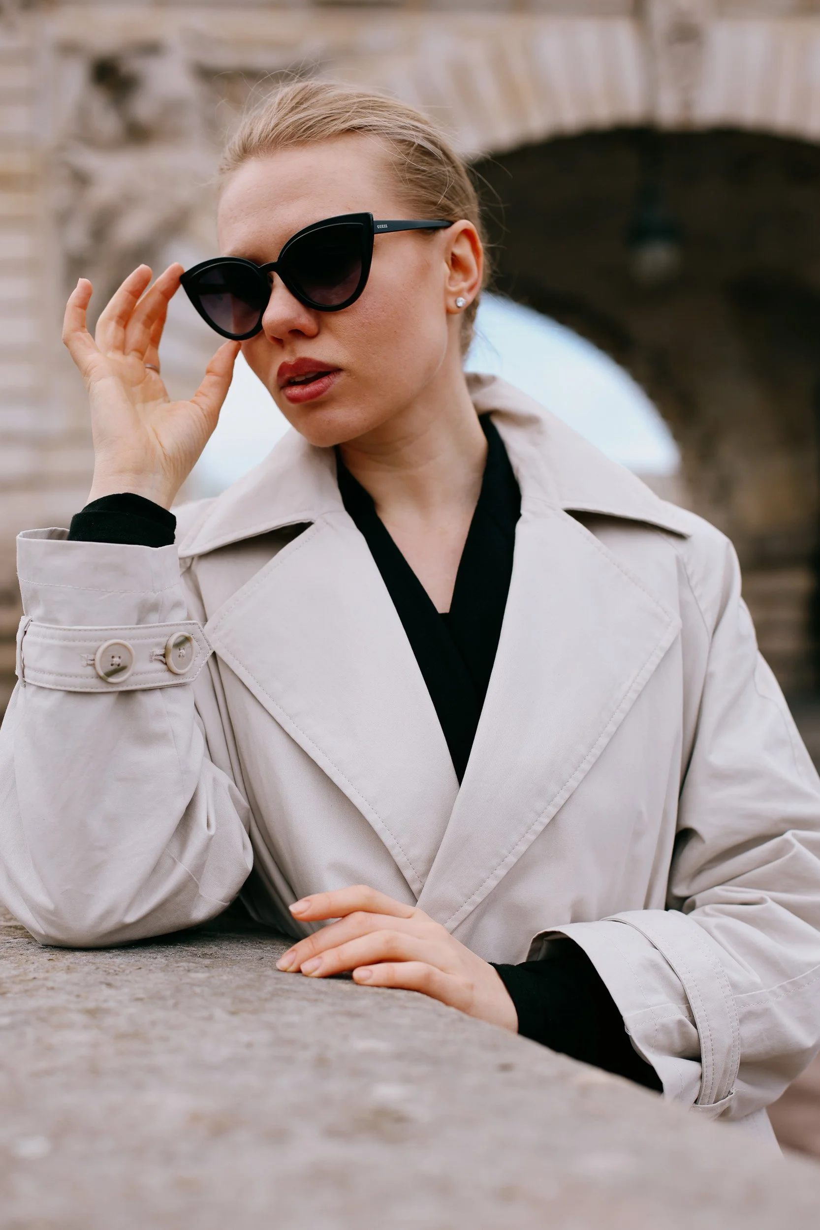 A woman with blonde hair wearing black sunglasses, a beige trench coat, and a black shirt, leaning on a concrete surface with an arched brick bridge in the background.