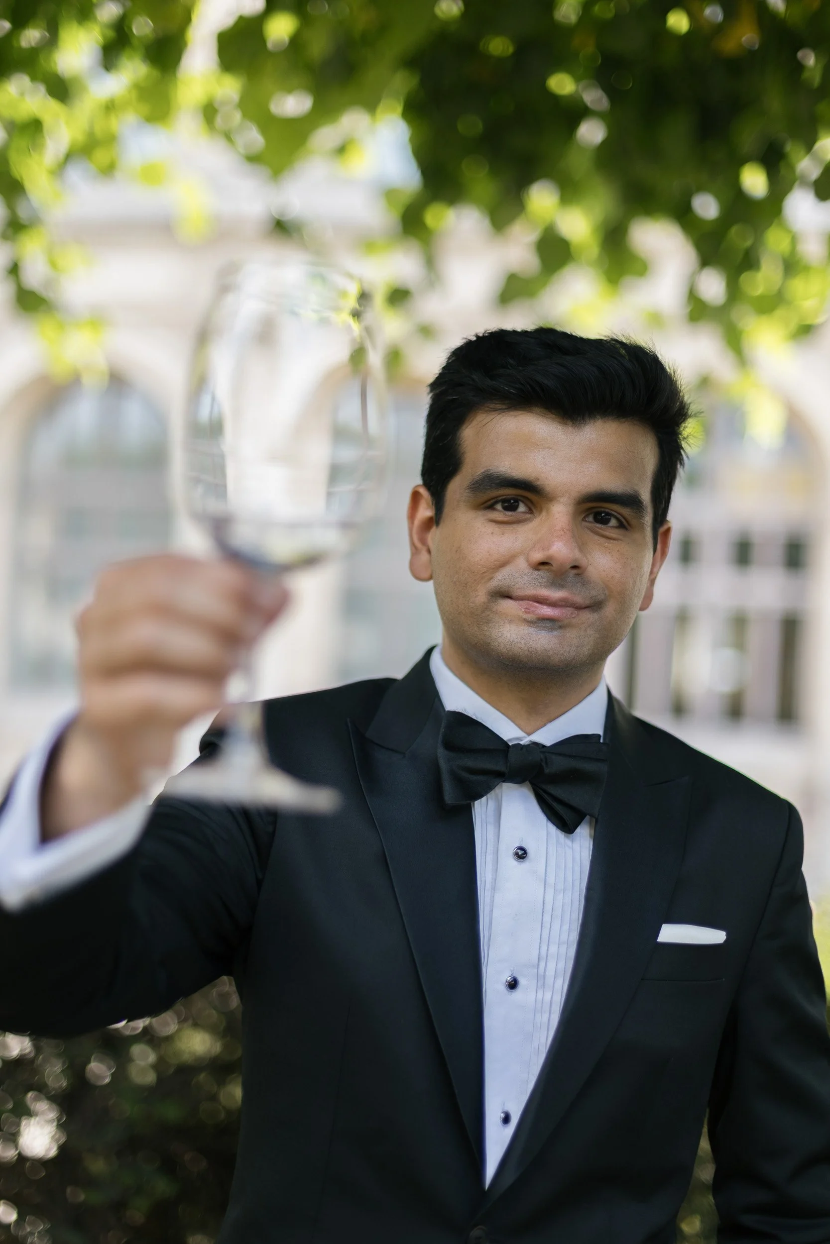 Man in a black tuxedo with a bow tie holding a glass of wine outdoors with green leaves overhead.