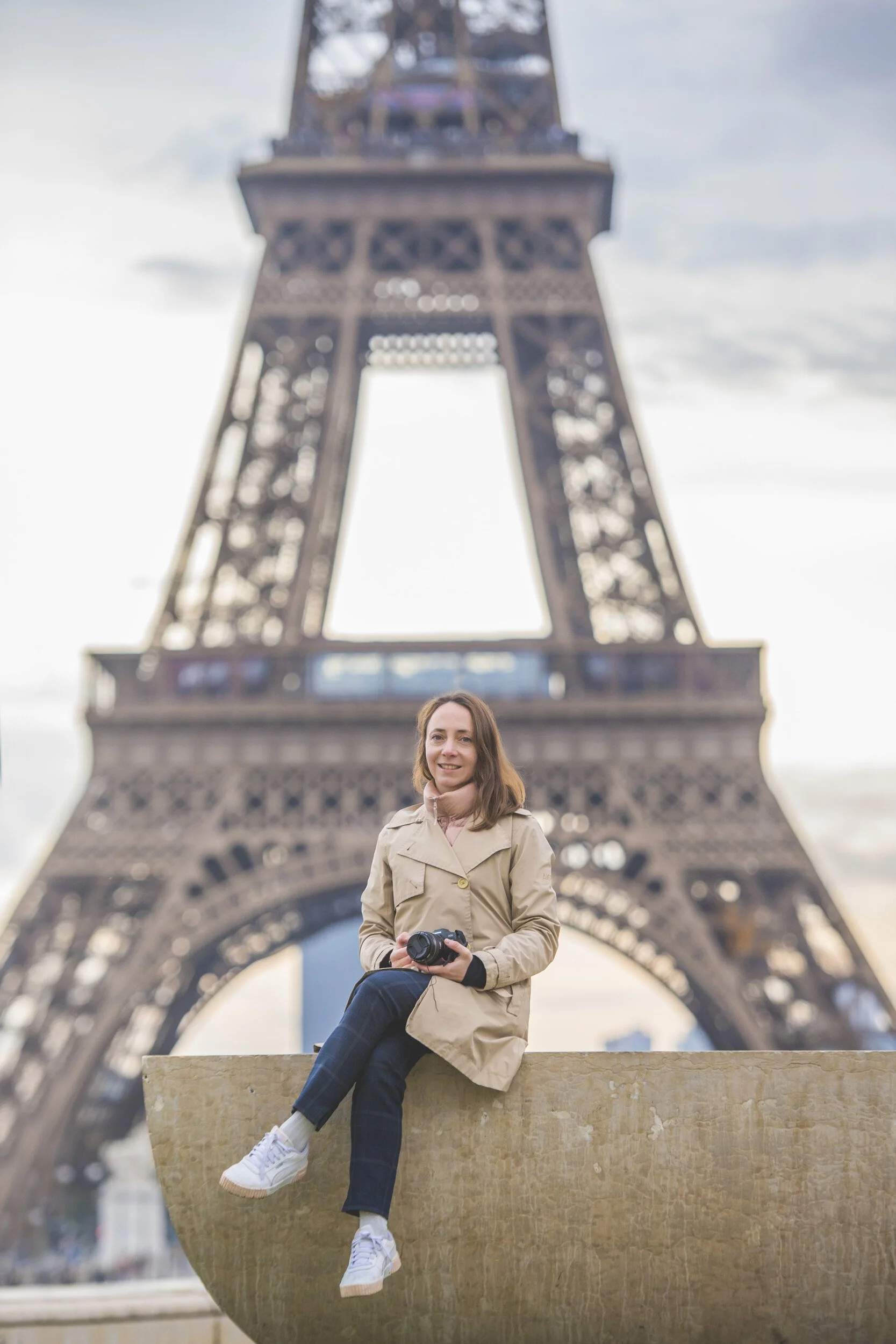 A woman sitting on a marble fountain in front of the Eiffel Tower, holding a camera, with the tower and a cloudy sky in the background.