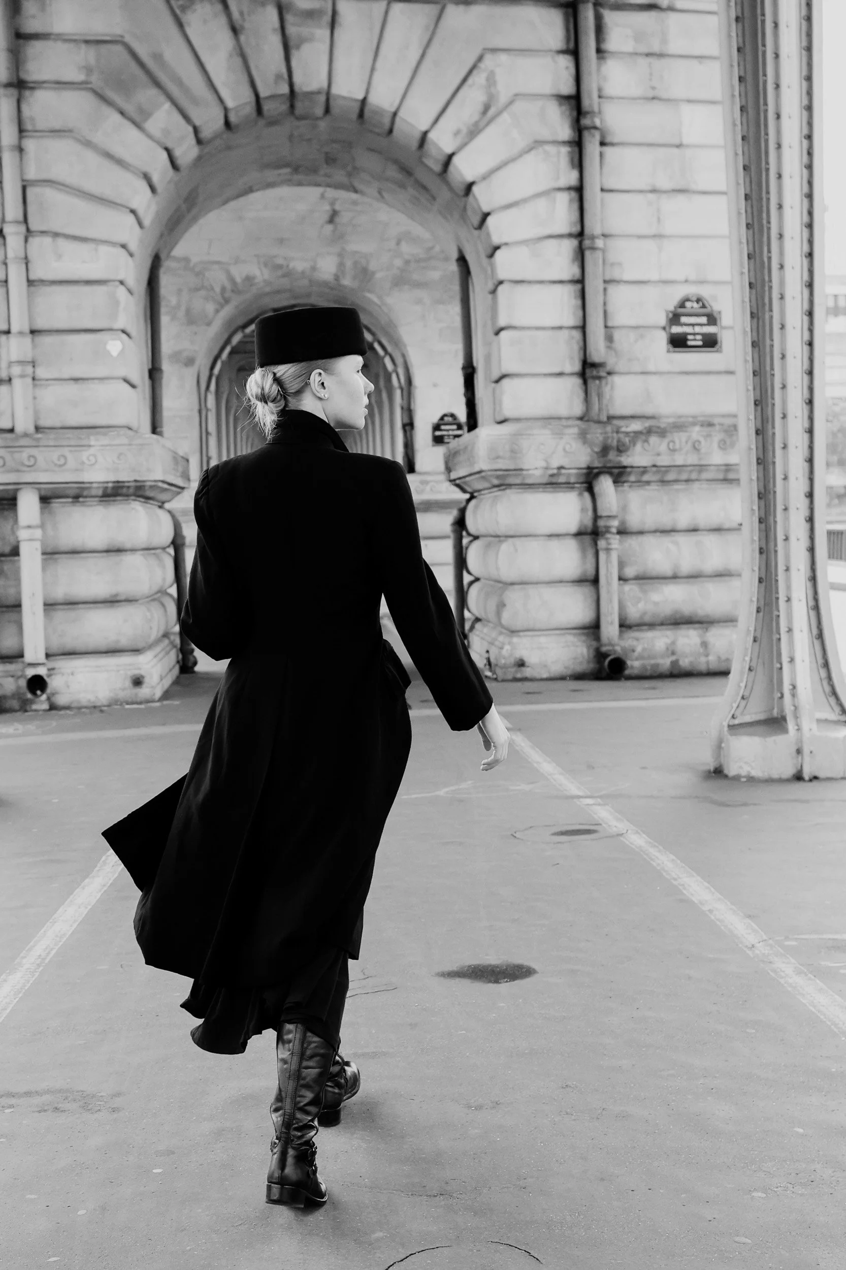 A woman in a black coat, hat, and knee-high leather boots walking in front of a stone archway in an urban setting, in black and white.