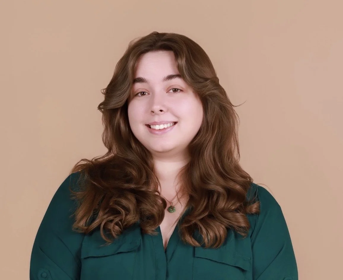 A woman with long wavy brown hair, smiling, wearing a green top and a necklace, standing against a beige background.