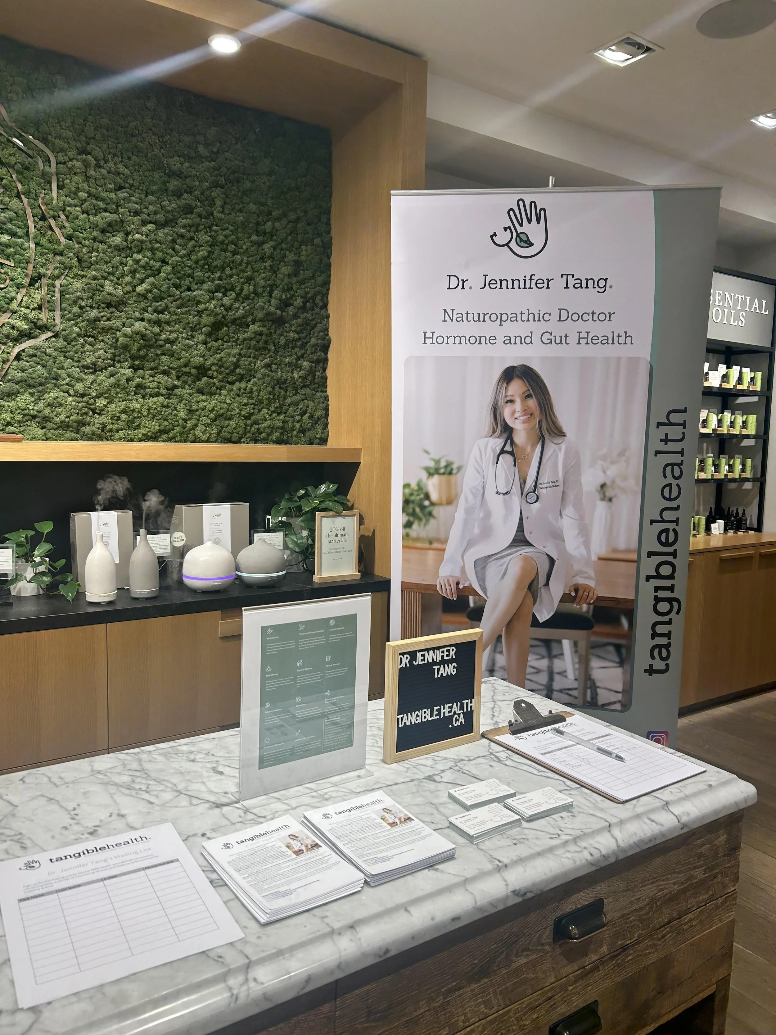 A promotional display for Dr. Jennifer Tang, a naturopathic doctor specializing in hormone and gut health. The display includes a large poster with her photo and information, a marble counter with informational pamphlets, brochures, a clipboard with a sign-up sheet, and several small bottles and products arranged on shelves.