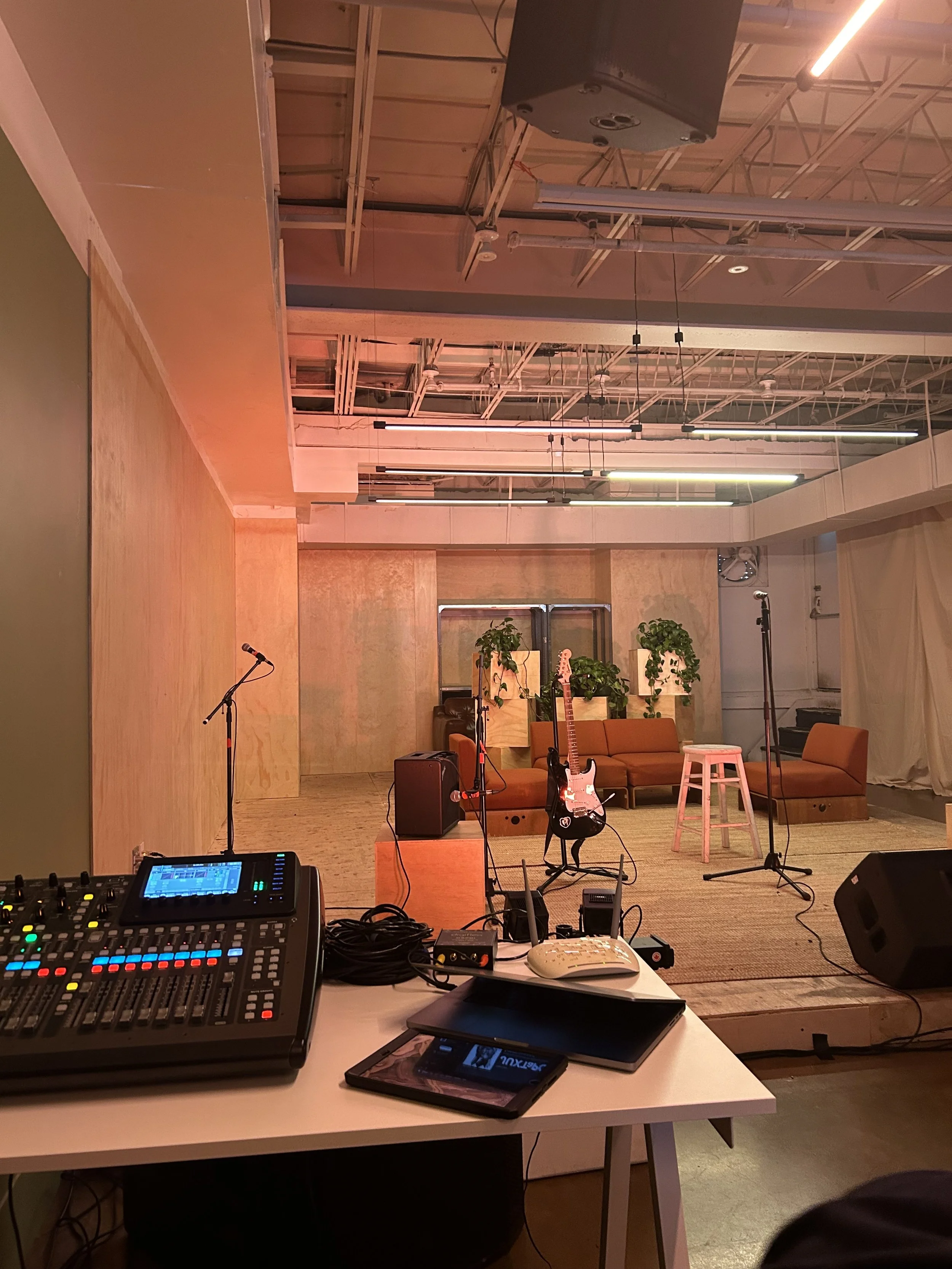 Photo of a performance space with musical equipment including microphones, guitars, and sound mixing console, set up in front of a seating area with chairs and plants, under a partially unfinished ceiling with exposed pipes and lighting.