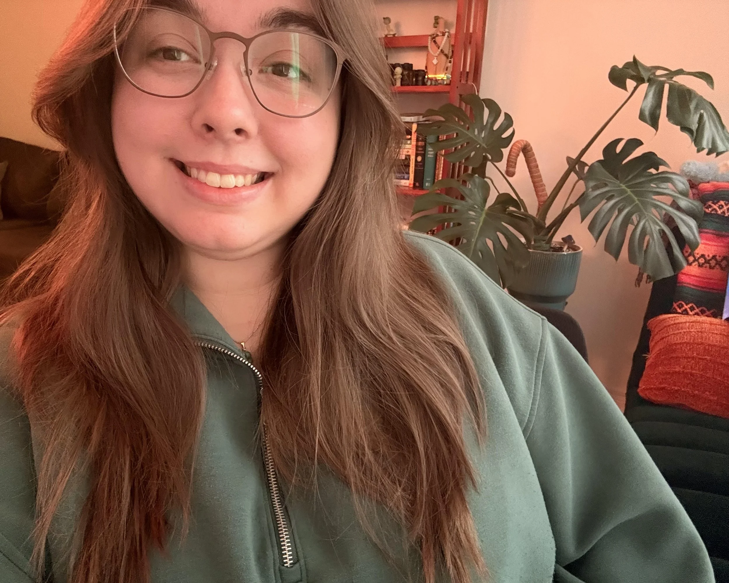 Close-up of a smiling woman with long red hair, wearing glasses and a green zip-up jacket, sitting indoors near a plant and a bookshelf.