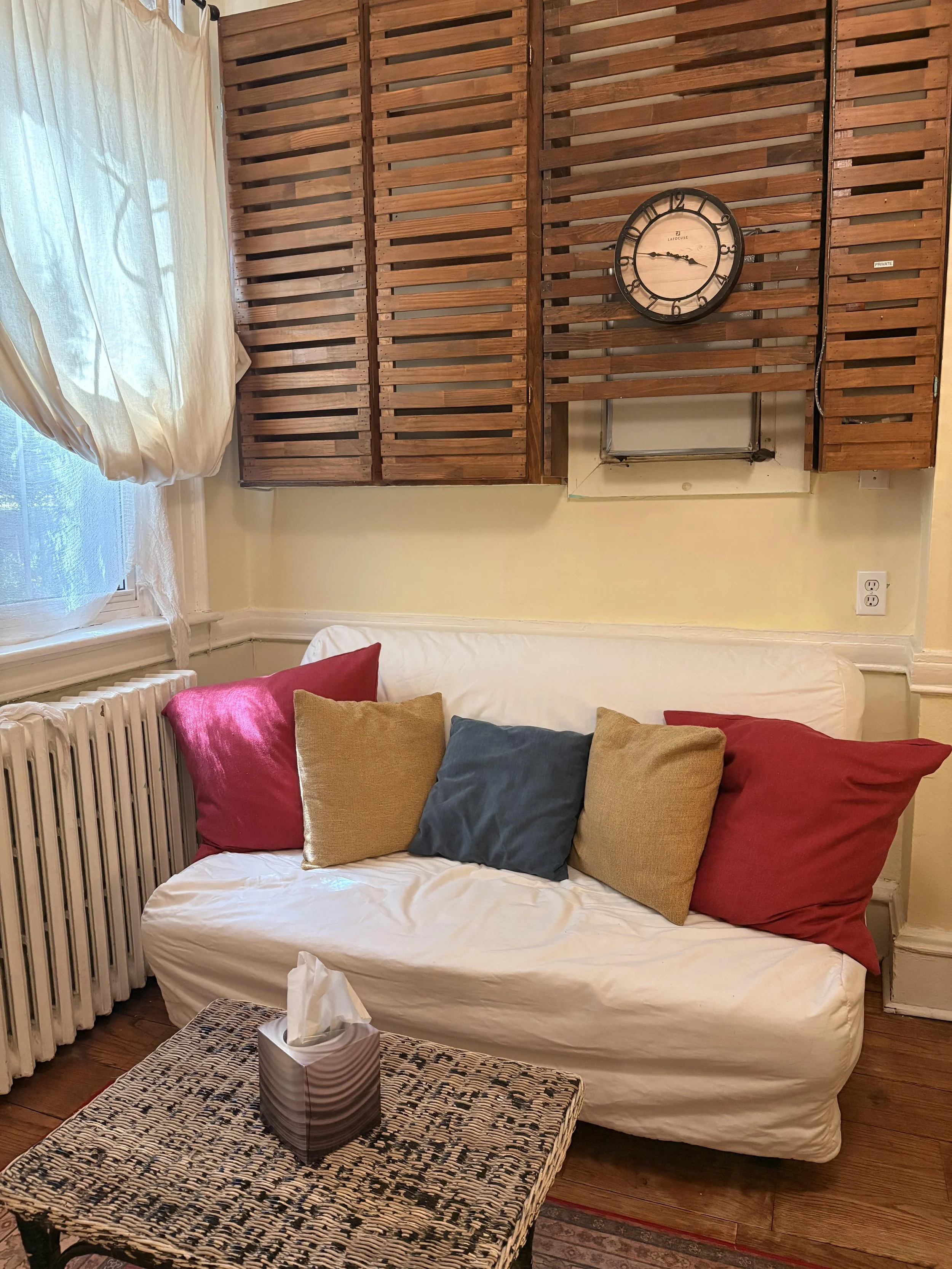 A cozy living room with a white sofa, colorful pillows, a woven coffee table with a tissue box on top, a window with white curtains, a wooden slatted wall, a clock, and a radiator.