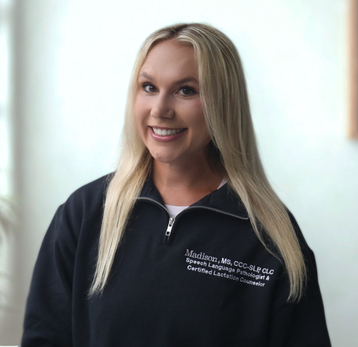A woman with long blonde hair smiling, wearing a black jacket with her professional credentials embroidered on it.