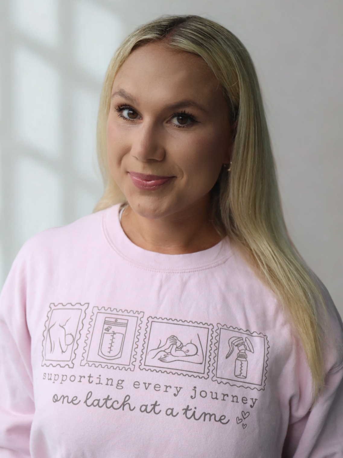 A woman with blonde hair wearing a pink shirt with illustrations of medical items and text supporting breastfeeding.