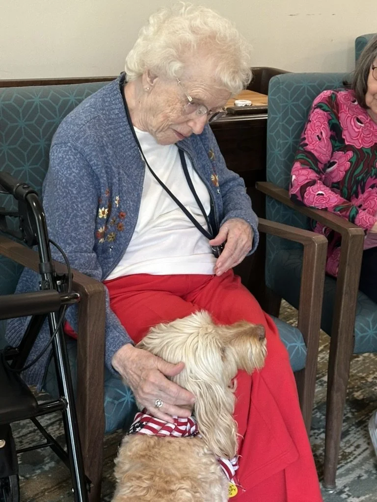 An elderly woman with white curly hair, glasses, and a blue cardigan is sitting in a chair, looking at and petting a small, light-colored dog wearing a red and white striped bandana. She is dressed in red pants and a white shirt, and there is another