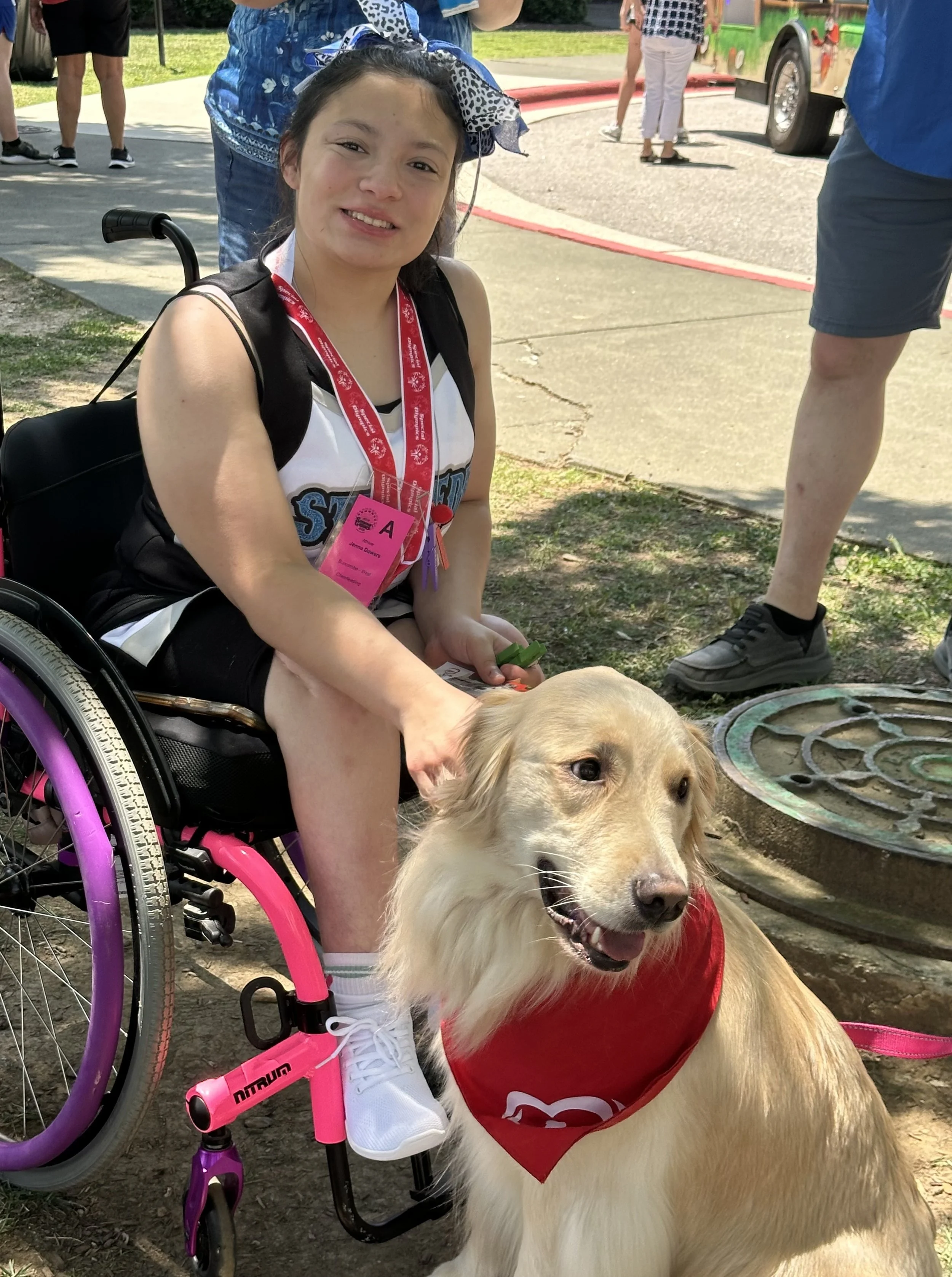 A young woman with a disability, sitting in a pink wheelchair, smiling and petting a golden retriever dog wearing a red bandana. The scene appears to be outdoors with people and a bus in the background.