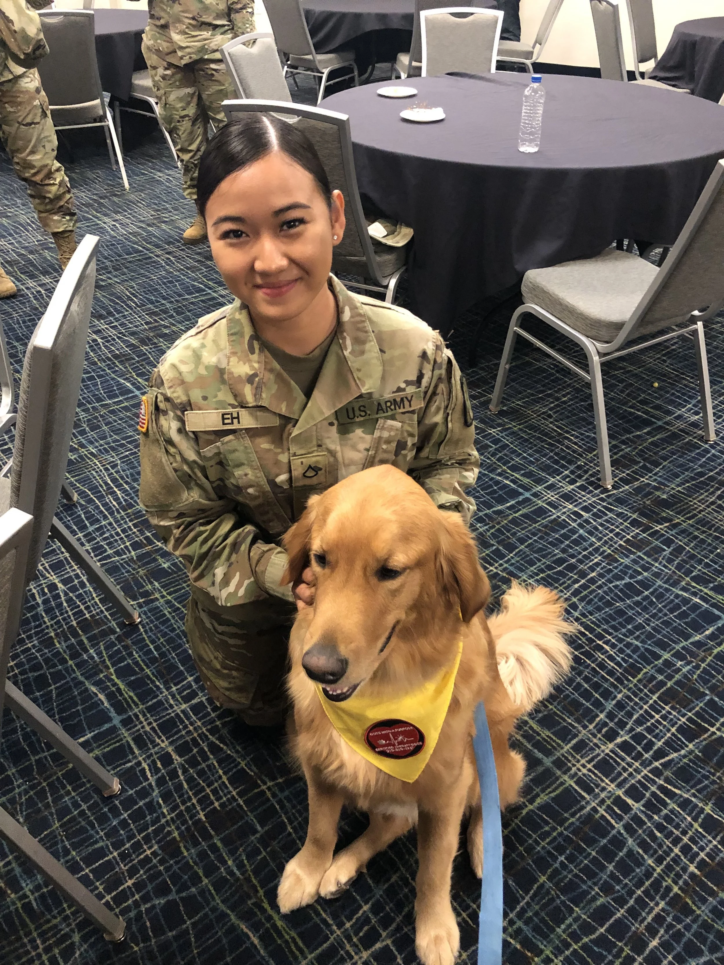 A smiling woman in U.S. Army camouflage uniform kneeling next to a golden retriever wearing a yellow vest with a red circular badge.