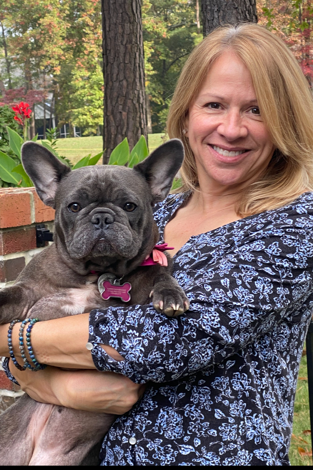 A woman with blonde hair smiling while holding a French Bulldog puppy outdoors in a garden with trees and plants in the background.