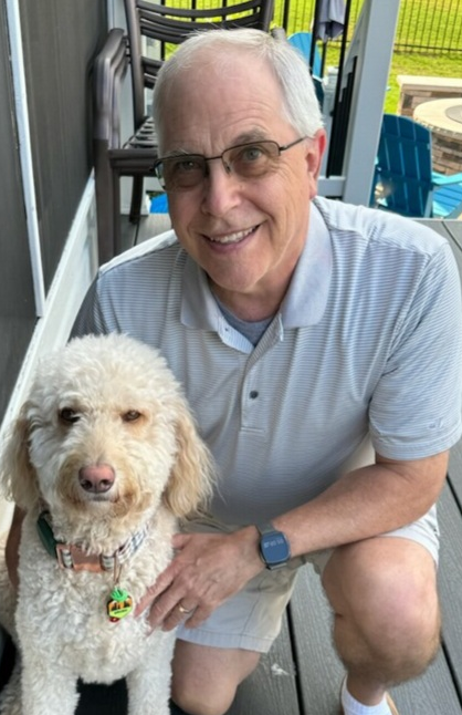 An elderly man with glasses smiling and crouching next to a white curly-haired dog on a porch with outdoor furniture and a grassy yard in the background.