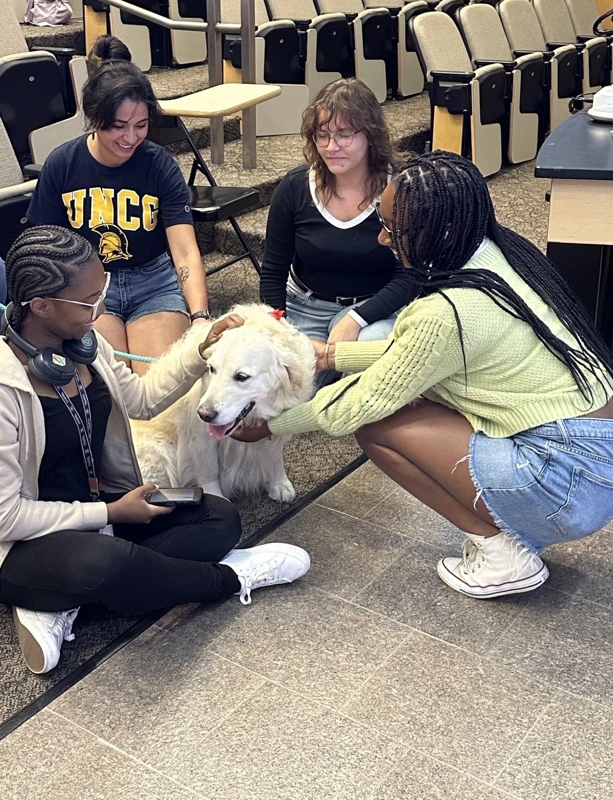 Four women and a service dog in a classroom or auditorium. One woman is sitting with headphones and a phone, petting the dog. Two women are holding and petting the dog's head and neck, while the fourth woman, with glasses and curly hair, looks on. Th