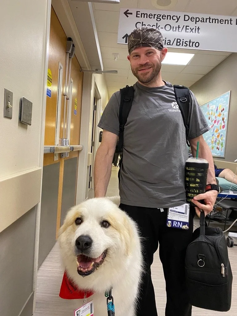 A man with a backpack and a travel mug standing next to a happy large white dog wearing a red vest, in a hospital corridor.
