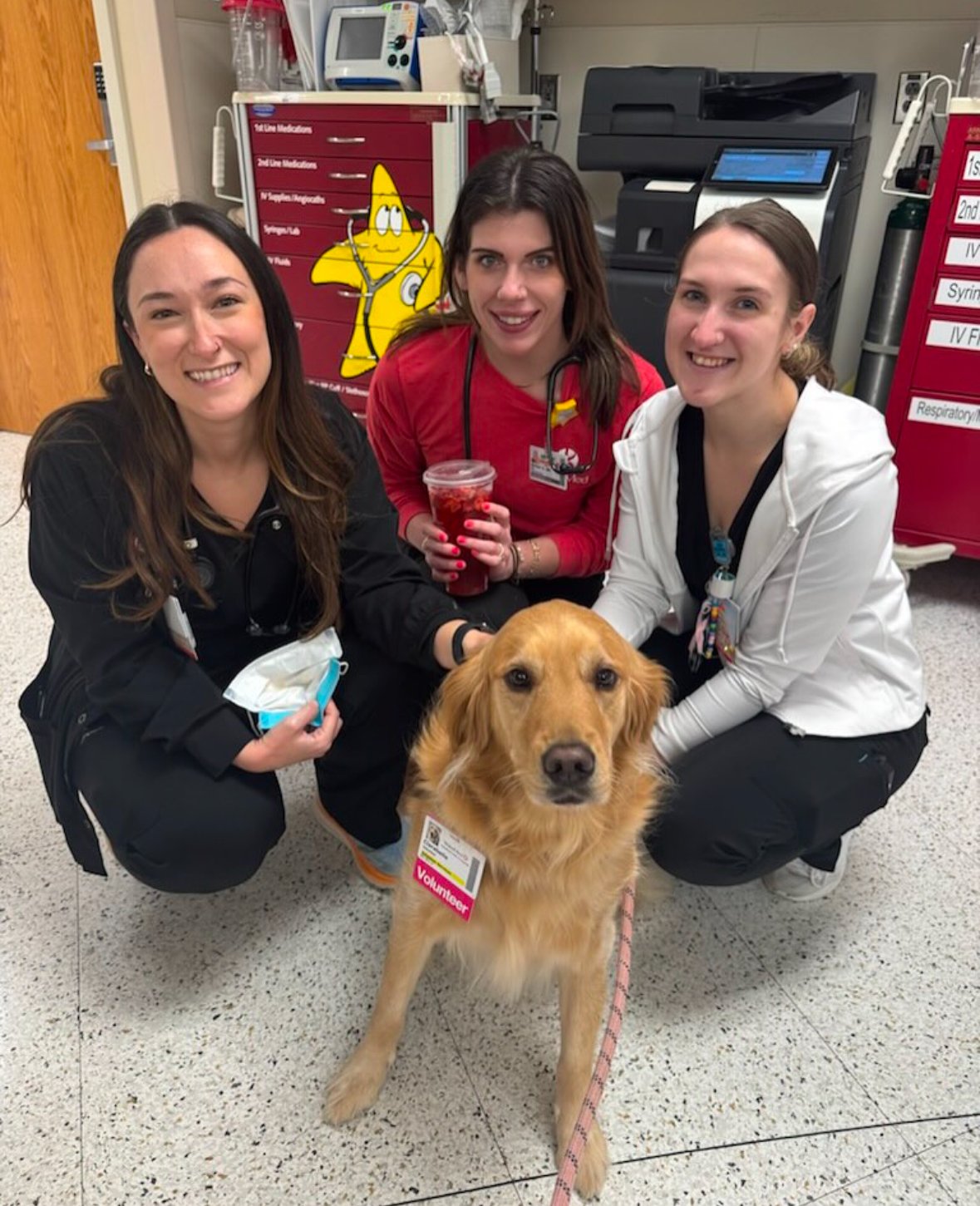 Three female healthcare workers and a volunteer dog in a hospital setting, with medical equipment and storage cabinets in the background. The dog is a golden retriever with a volunteer badge, sitting on the floor.