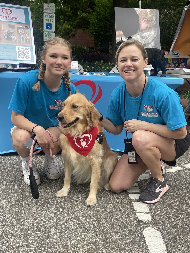 Two women in blue shirts are kneeling on either side of a golden retriever dog with a red bandana, at an outdoor event. There is a booth with informational posters and signs in the background, along with trees and a parking sign.