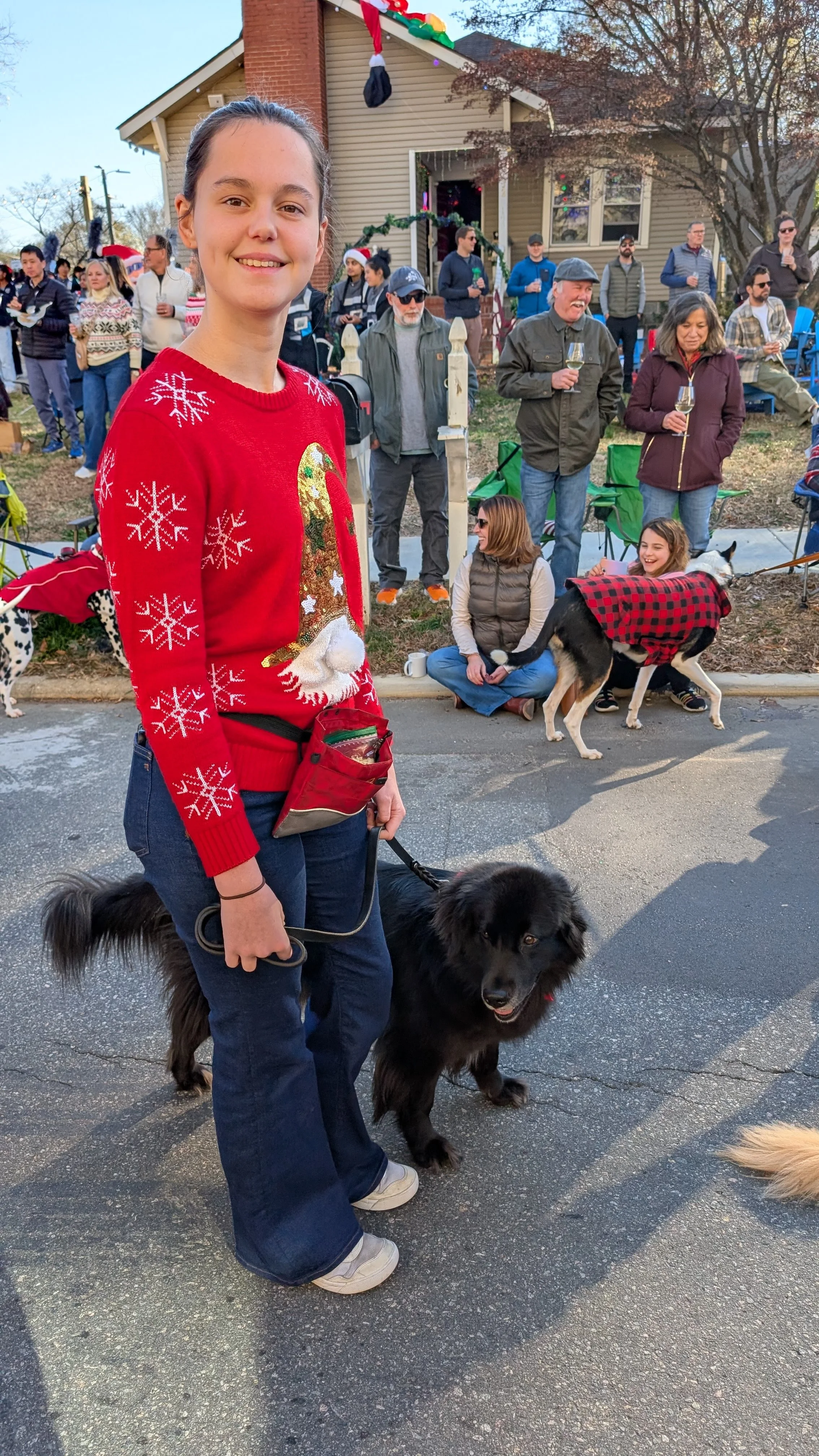 A young woman with a dog at a festive outdoor gathering, wearing a Christmas sweater with a gingerbread man and snowflakes, smiling at the camera. In the background, people are gathered outside a house decorated for the holiday season.